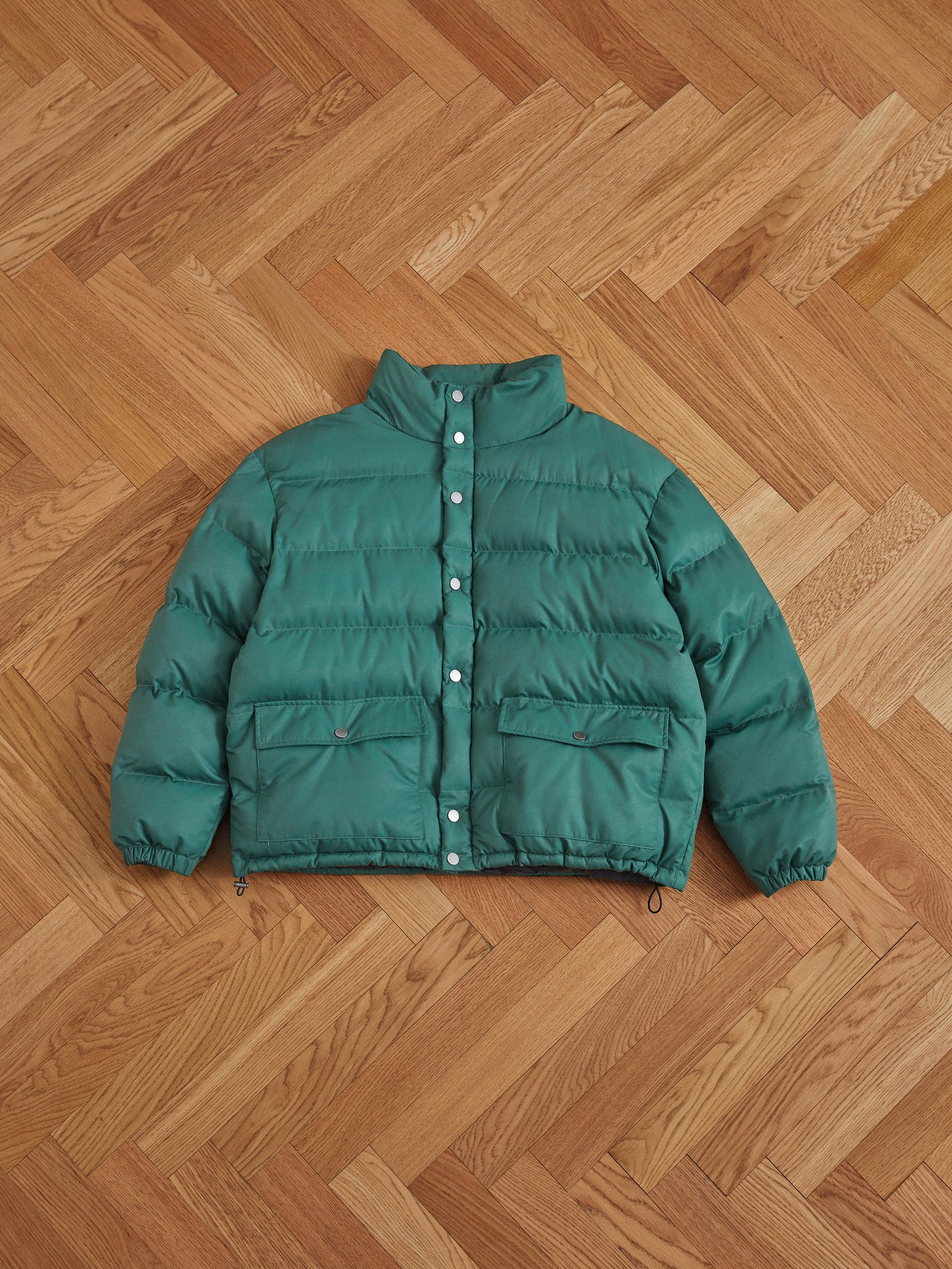 A deep green Found Laurel Pine Puffer Jacket with pockets laying on a wooden floor.