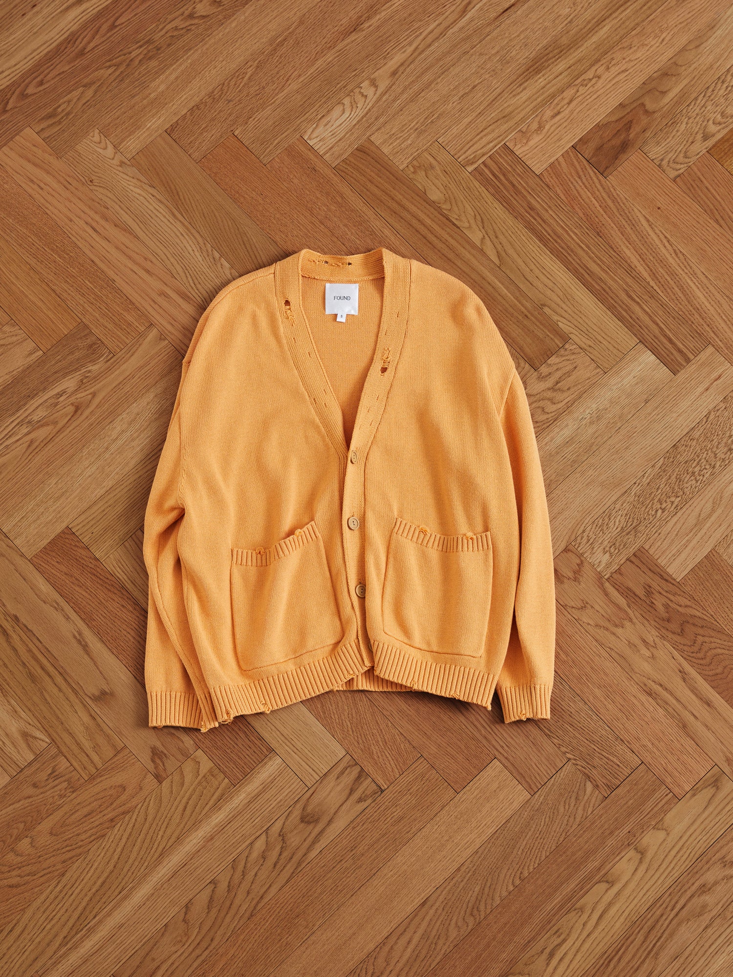 A Cadmium Distressed Cardigan sweater resting on a distressed wooden floor.