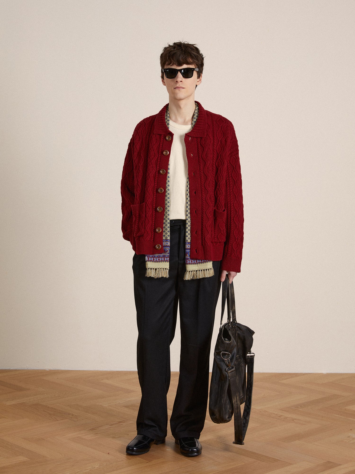 A man wearing sunglasses and a red hued yarn Parsidan Cable Knit cardigan from Found holding a bag.