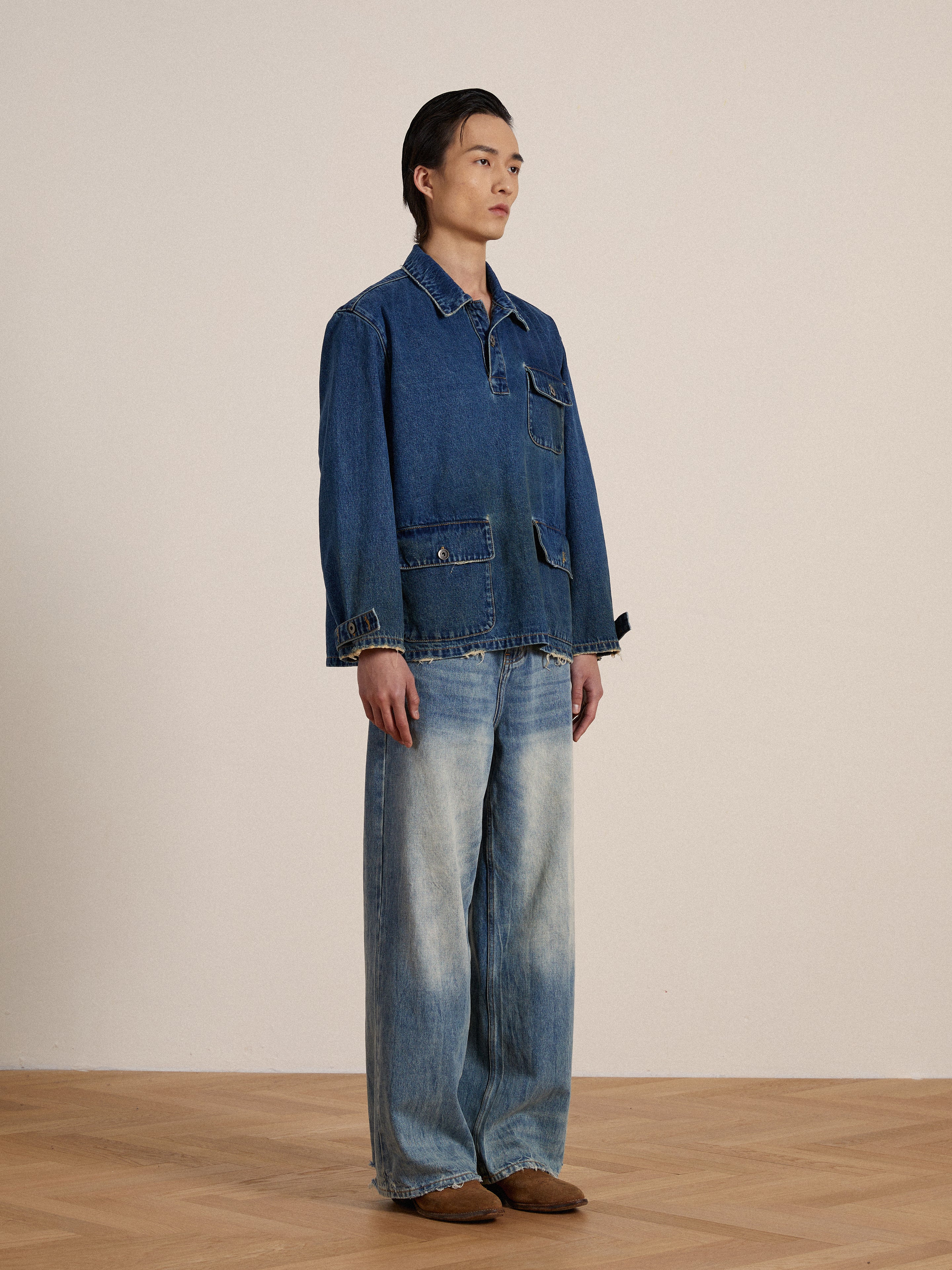 A man stands against a neutral background wearing a Found Azin Denim Pullover Work Shirt and jeans, paired with brown shoes, looking to the side.
