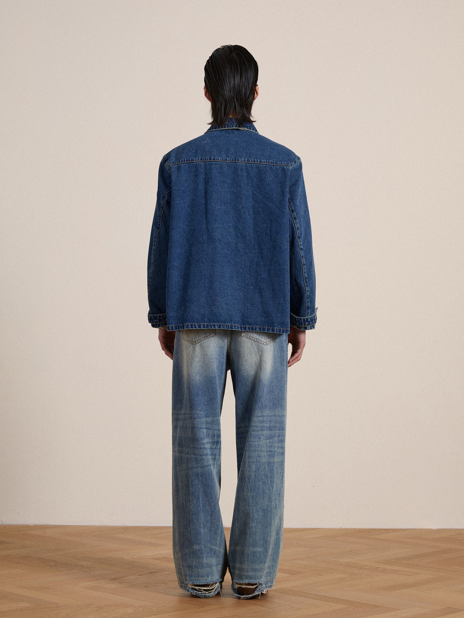 Rear view of a person wearing a Found Azin denim pullover work shirt and wide-legged jeans, standing in a room with a beige wall and wooden floor.