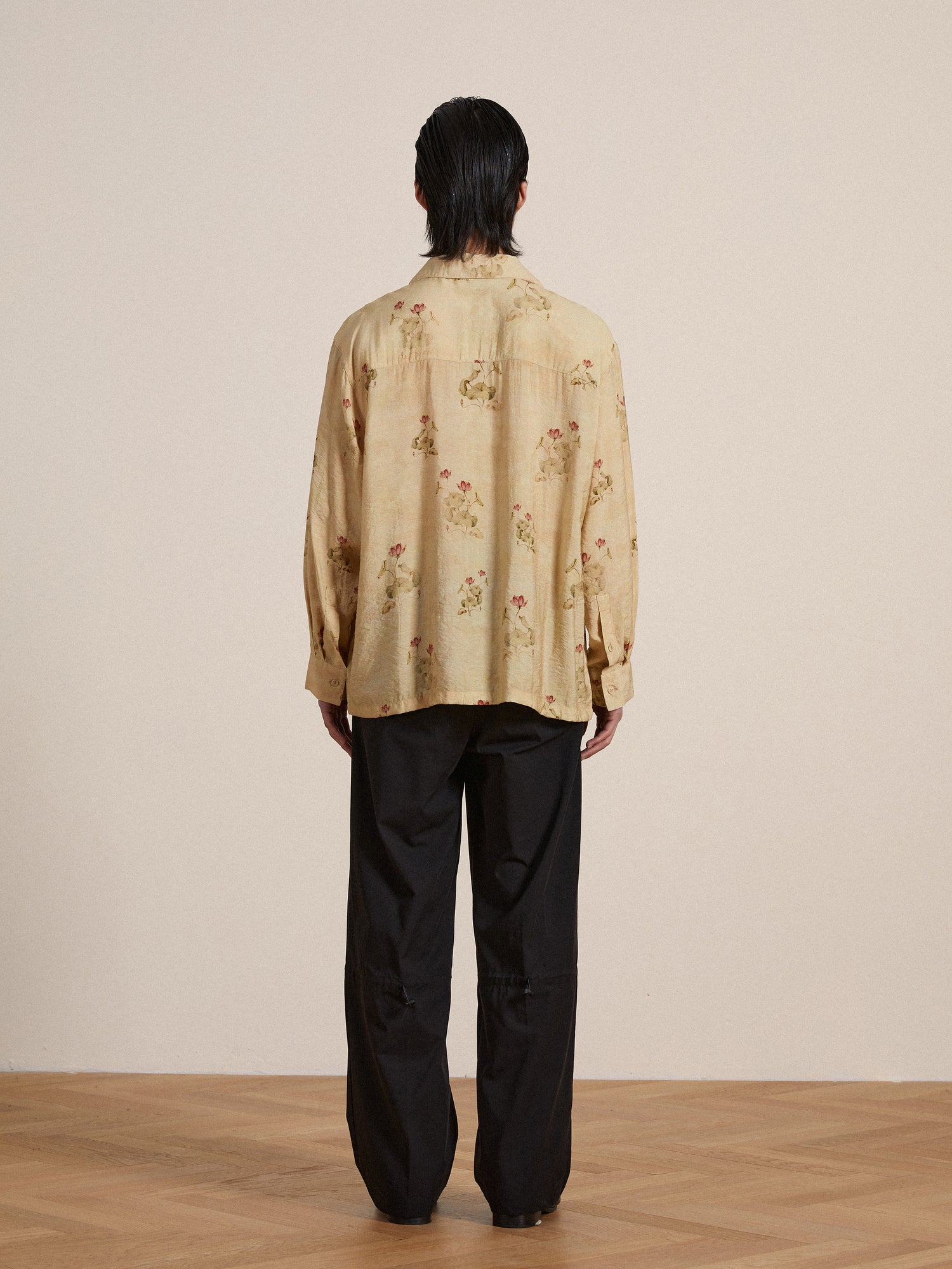 A person viewed from behind, wearing a beige floral shirt and black Found Tencel Pleated Pants, standing against a plain wall on a wooden floor.