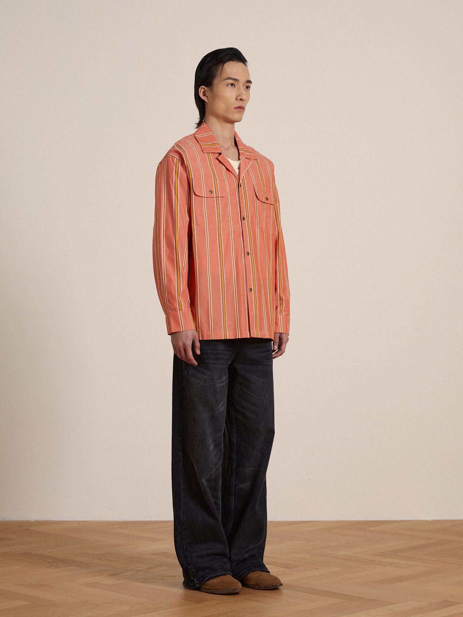 A man in an orange Found Stripe Citrus LS Camp Shirt standing on a wooden floor, exuding classic appeal.