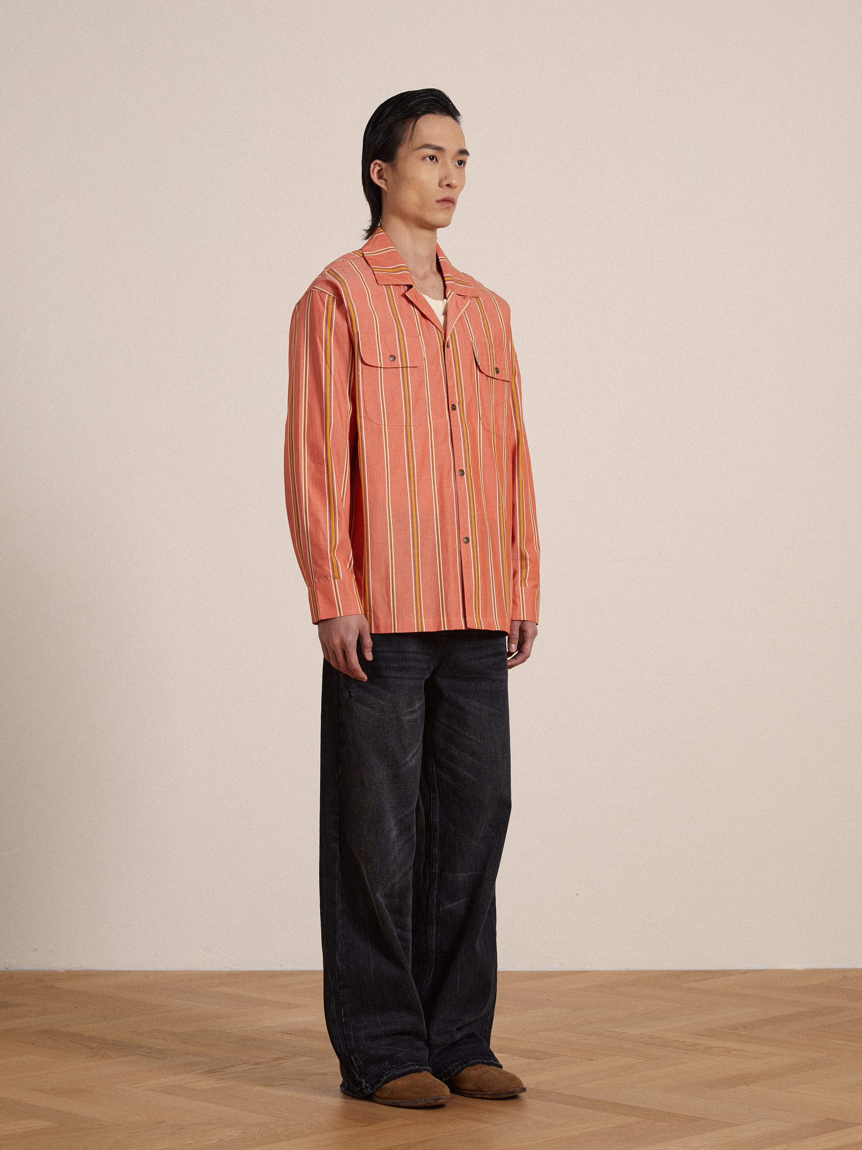A man in an orange Found Stripe Citrus LS Camp Shirt standing on a wooden floor, exuding classic appeal.