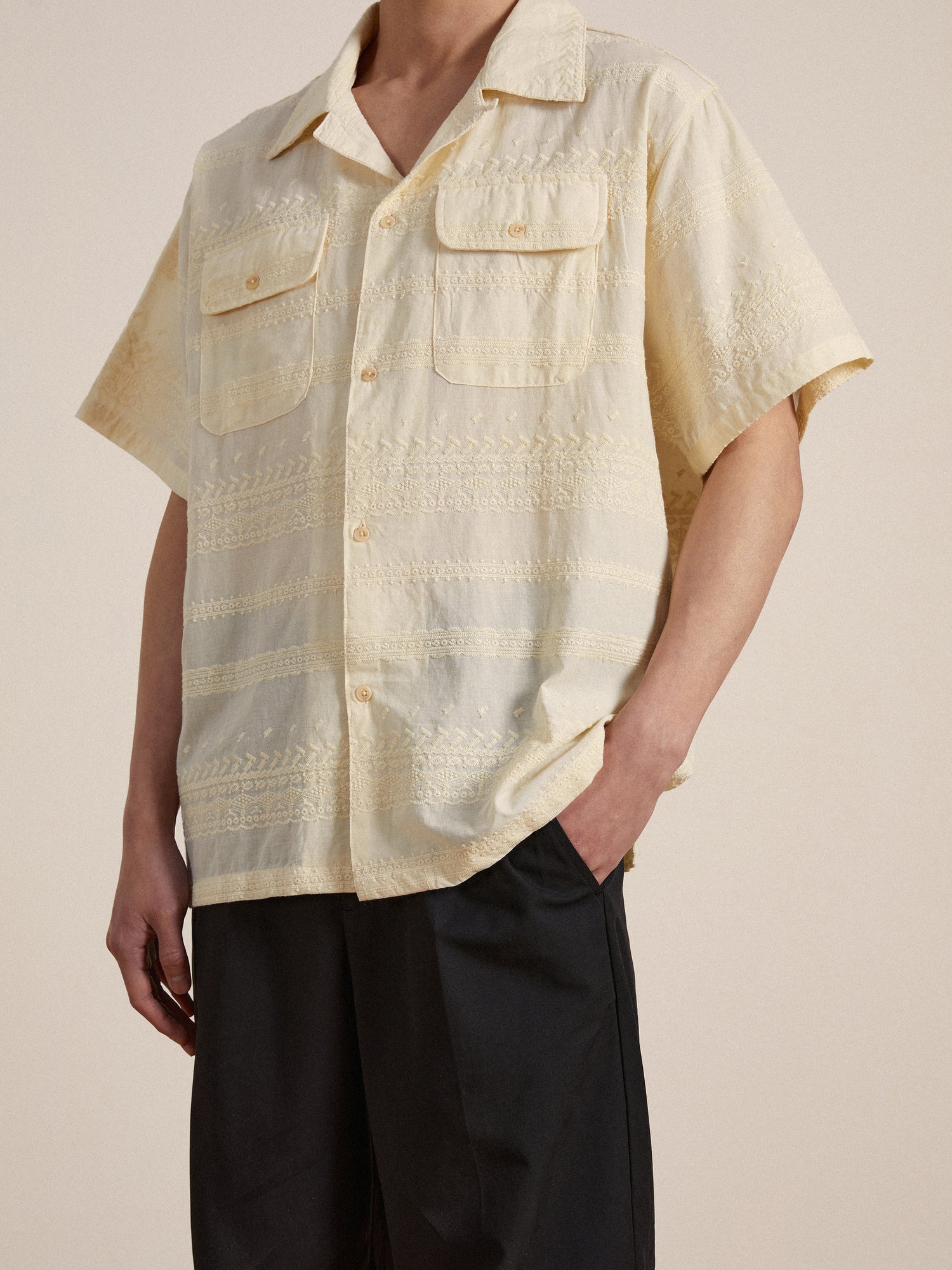 A man wearing a delicate lace detailing yellow Found Lace SS Camp Shirt and black pants.