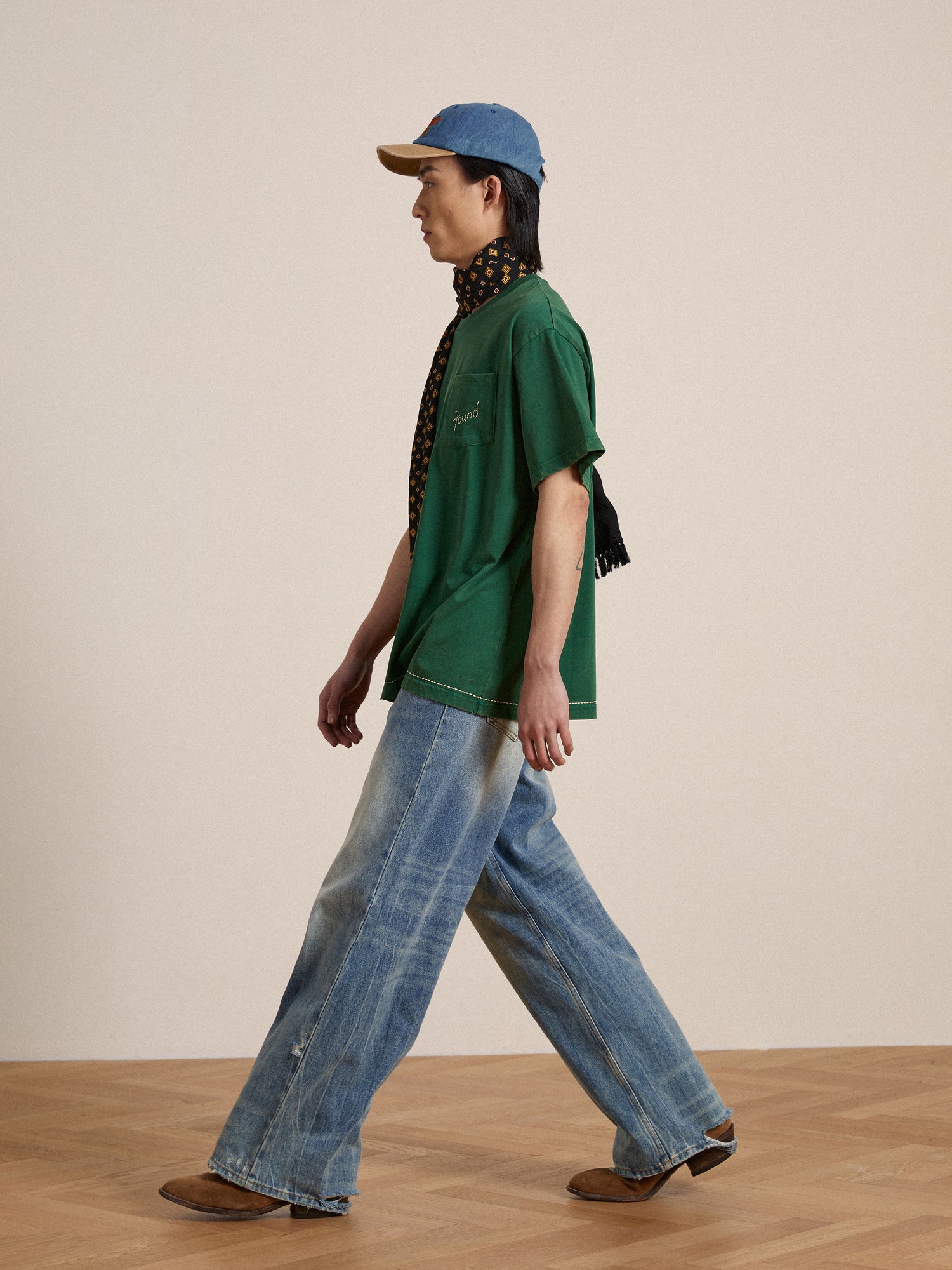 A man in a green Found Embroidered Logo Tee and blue baggy jeans walking on a wooden floor.
