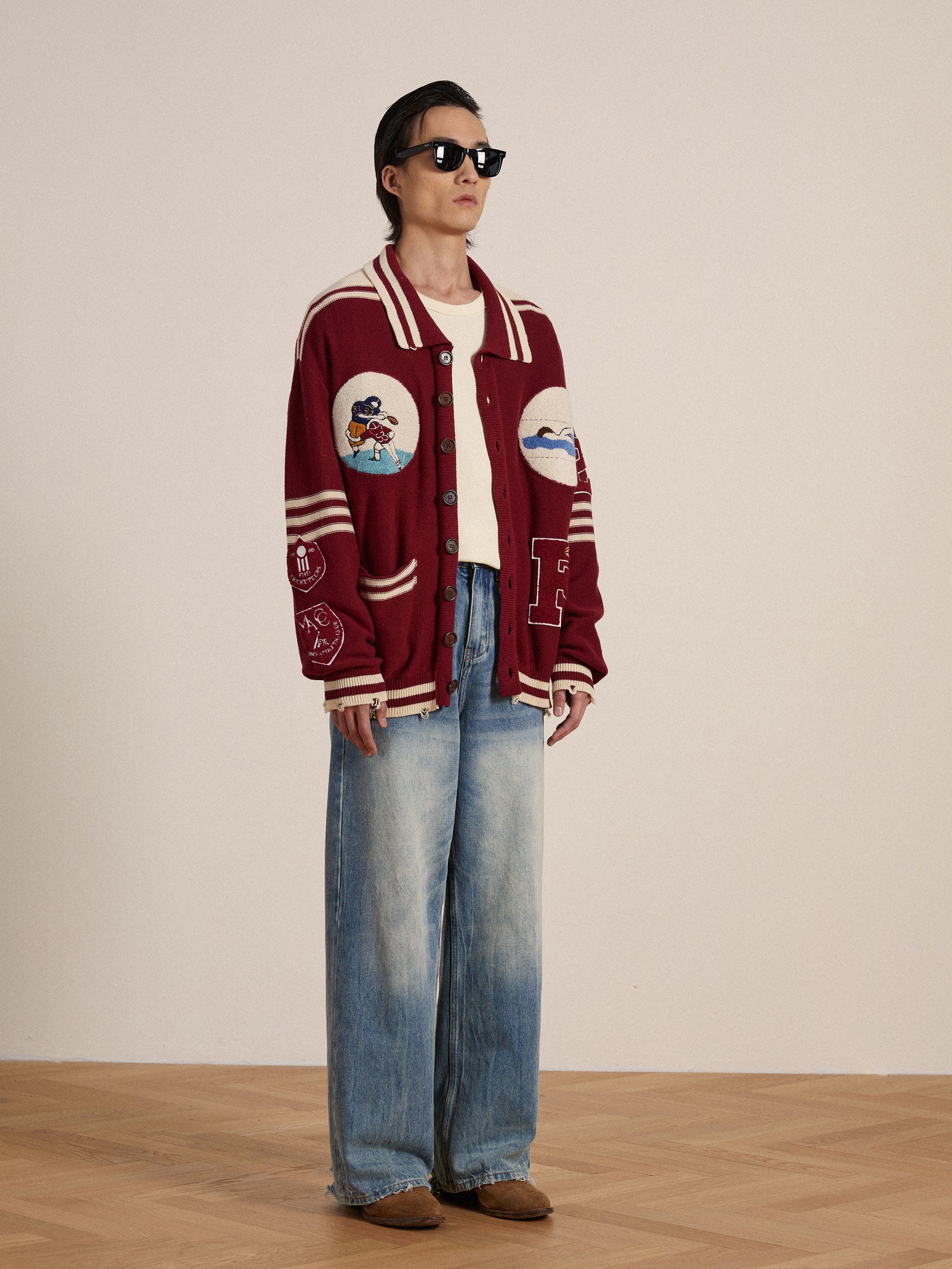 A woman in a red Found Varsity Patch Collared Cardigan and jeans is standing on a wooden floor.