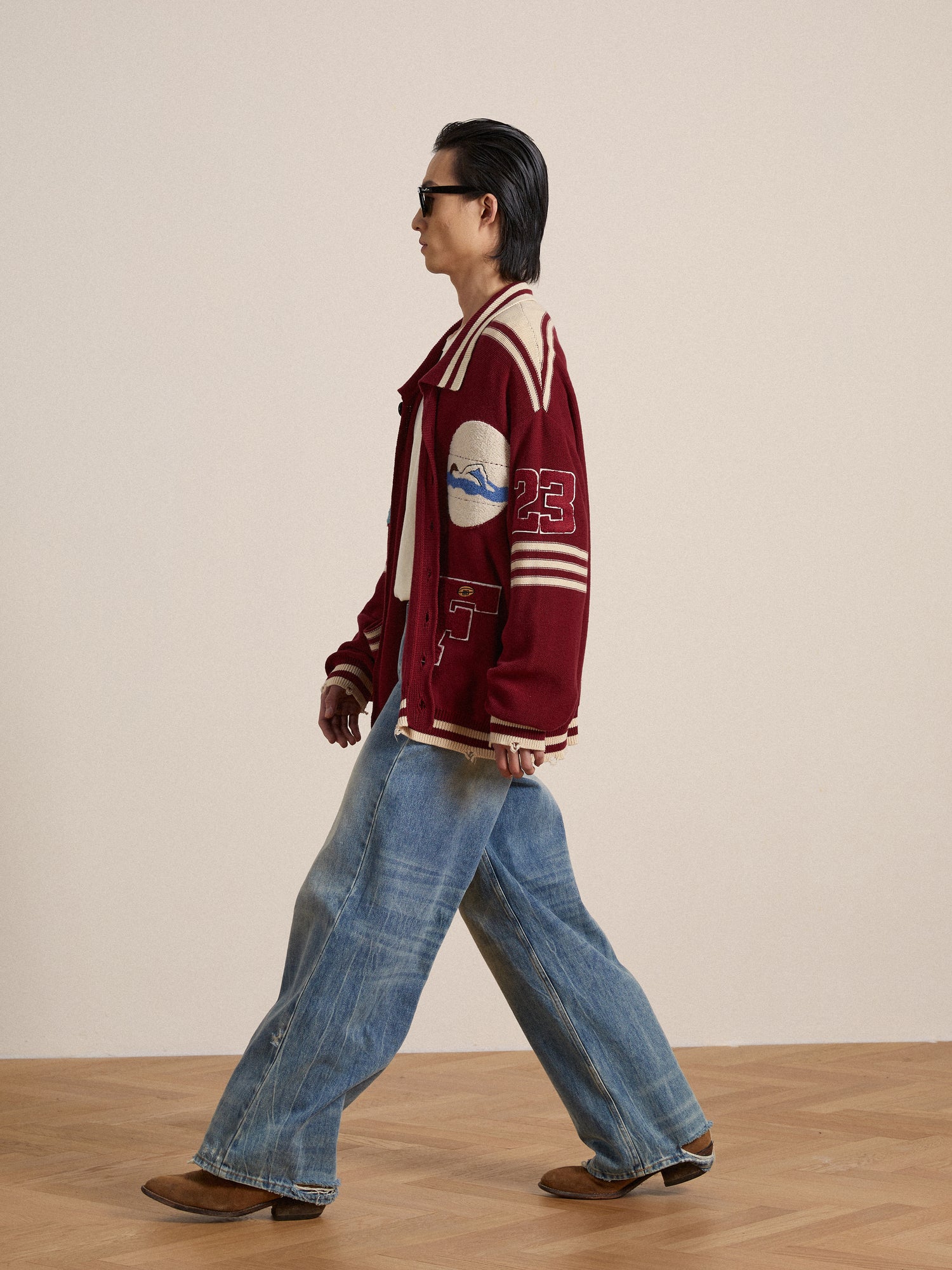 A man in a Found Fin Varsity Patch Collared Cardigan adorned with embroidered chenille patches and jeans walking on a wooden floor.