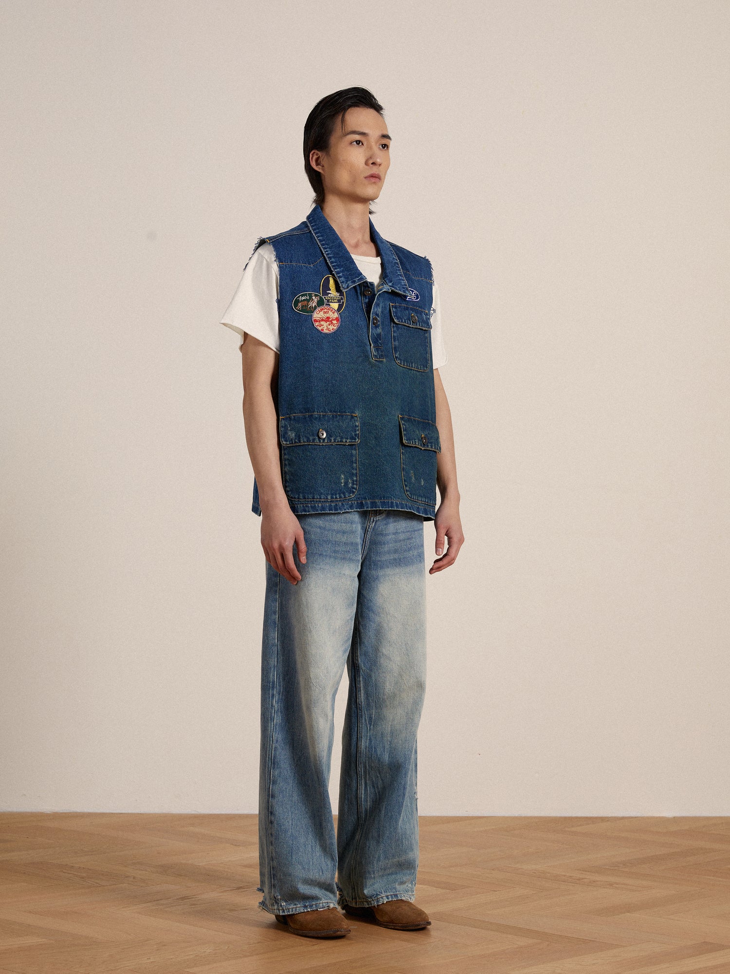A man in a vintage-inspired Found Raw Cut Patch Mechanic Denim vest adorned with embroidered patches, standing on a wooden floor.