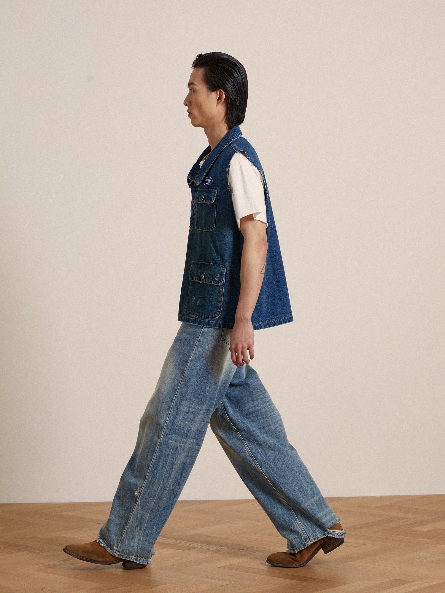A man in a Found Raw Cut Patch Mechanic Denim vest adorned with embroidered patches walking on a wooden floor.