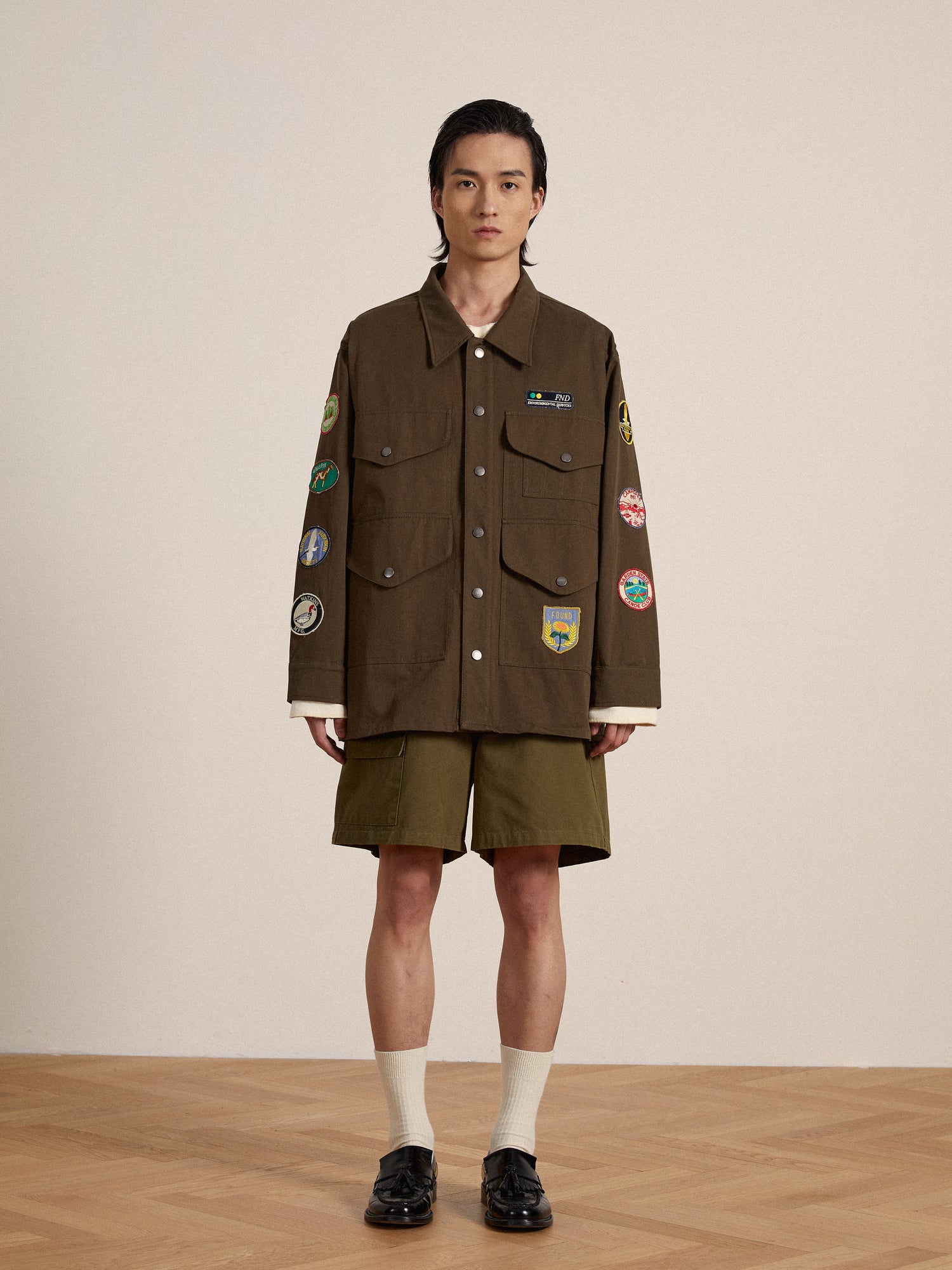 A man in a Found Ports Park Multi Patch Work Jacket and shorts standing on a wooden floor.