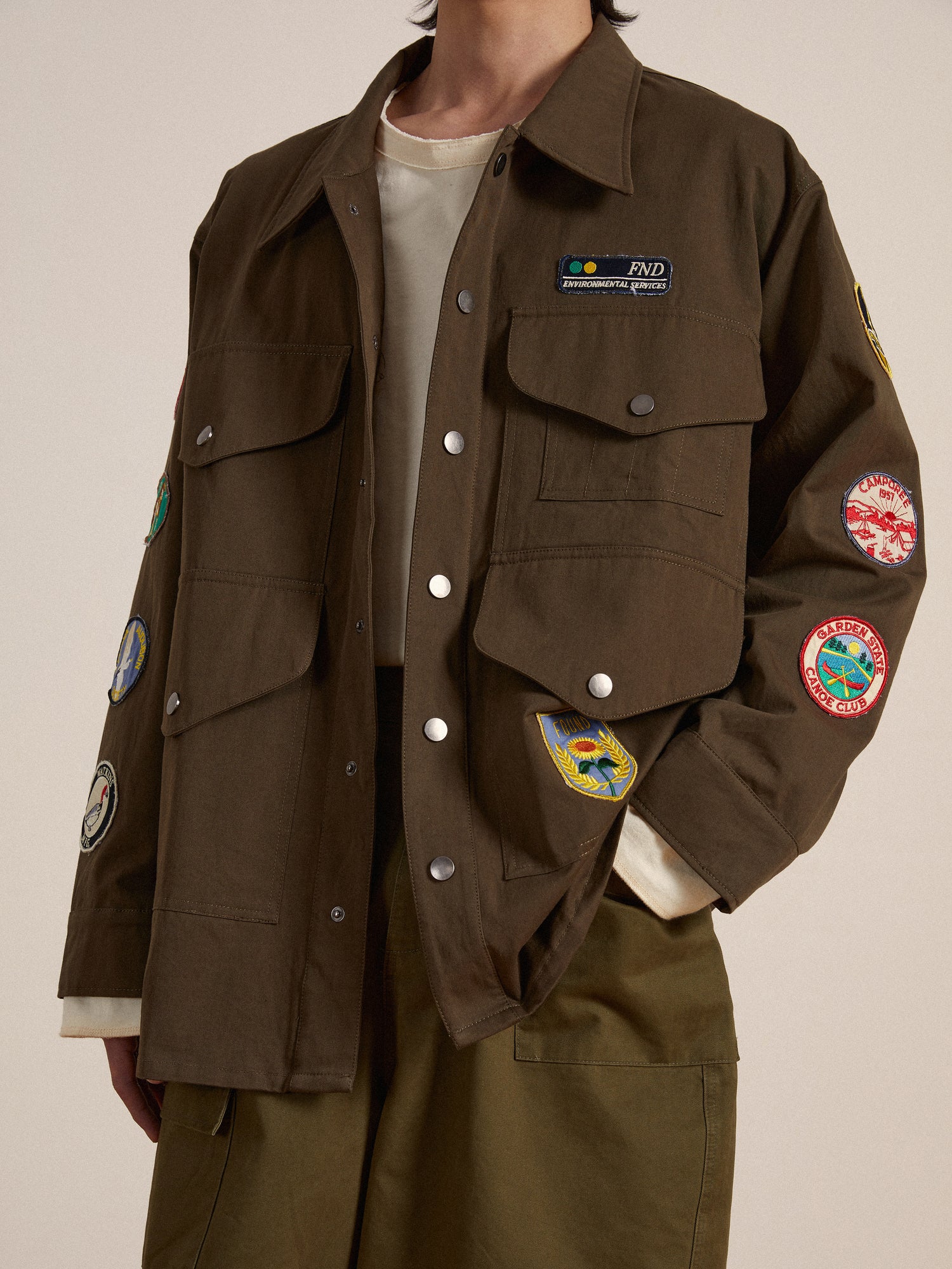 A man wearing a brown Found Ports Park Multi Patch Work Jacket with embroidered patches on it.
