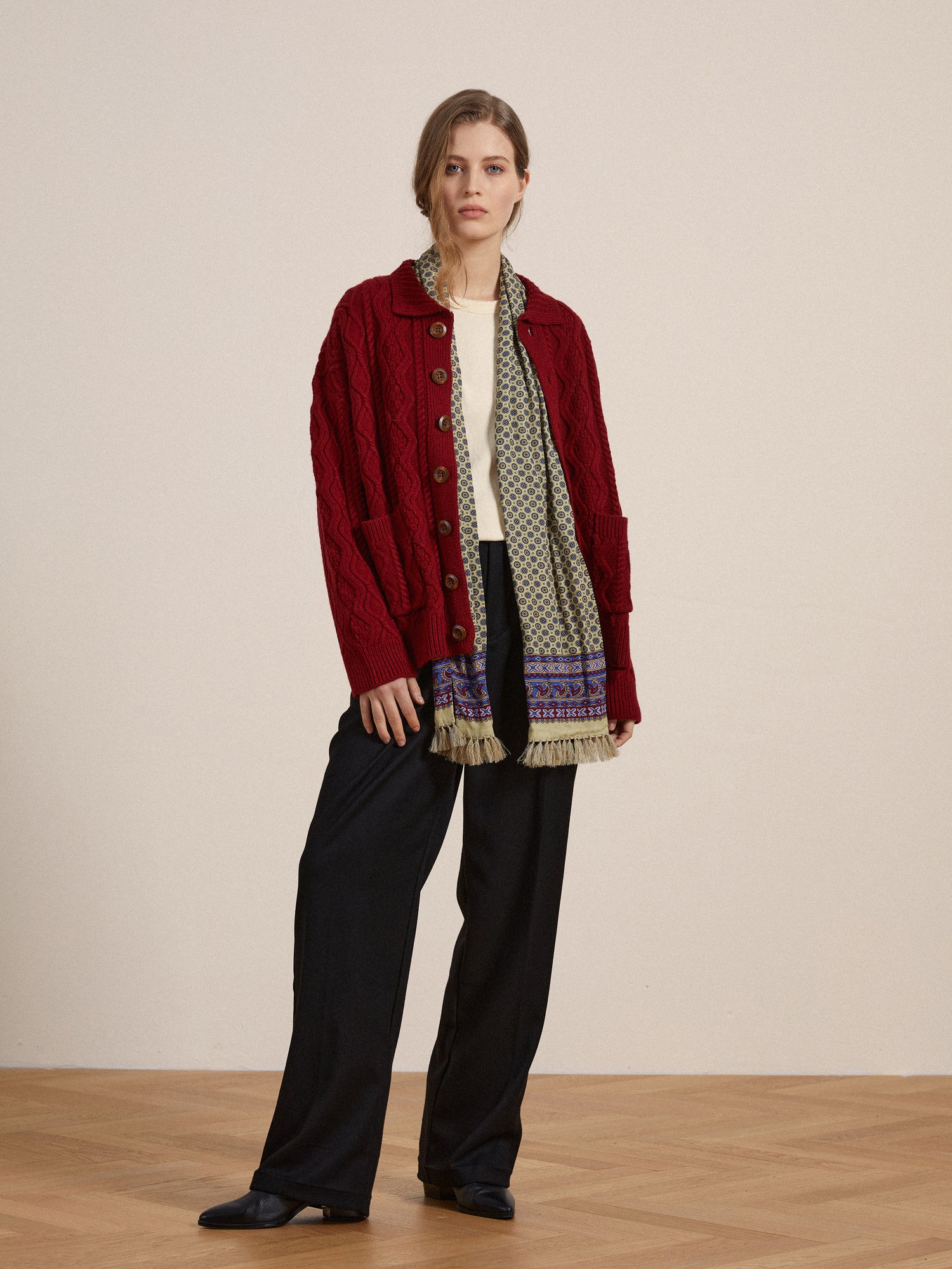 A woman wearing a Found Parsidan Cable Knit Cardigan in red hue and black trousers.