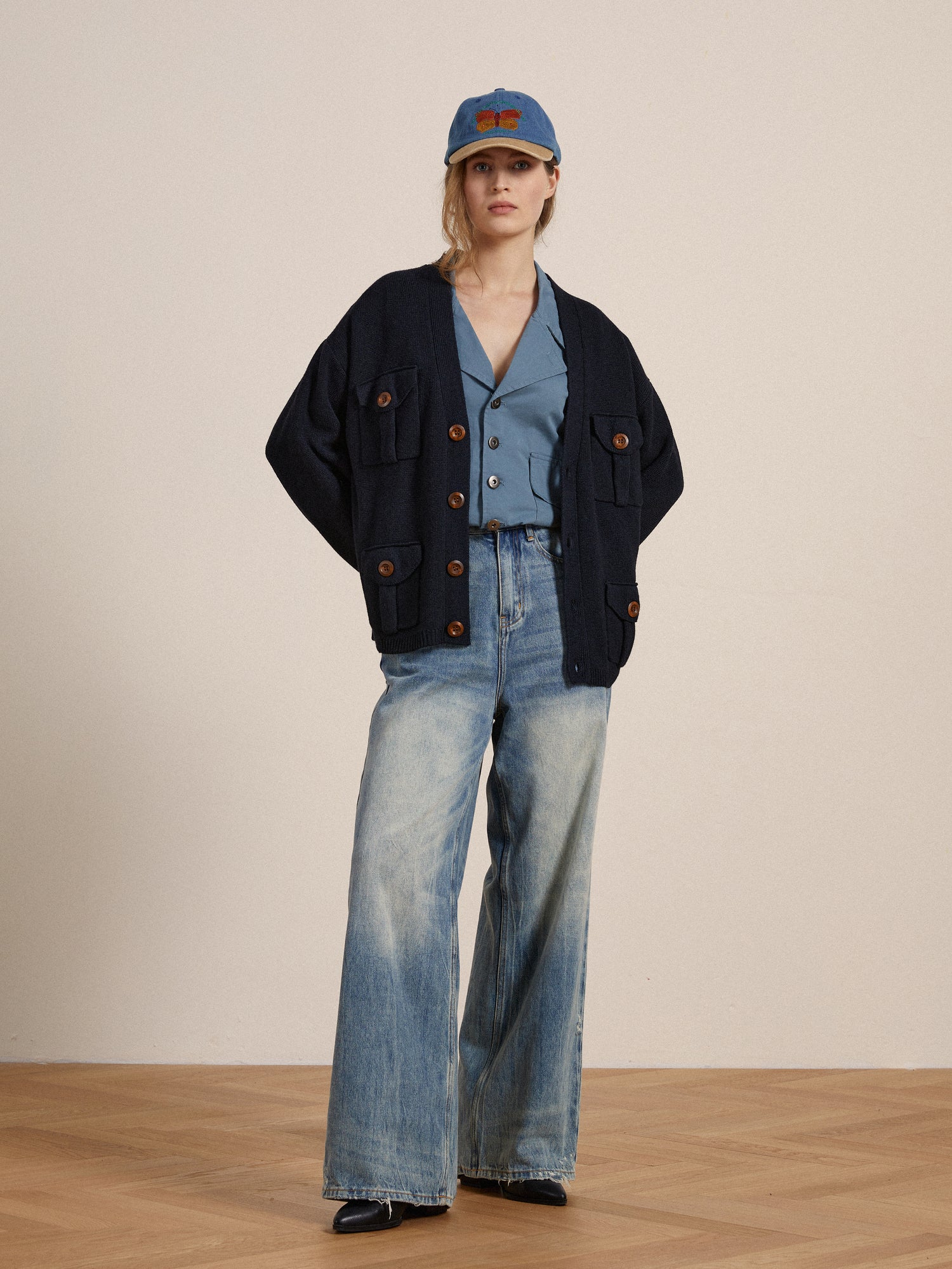 A woman in blue jeans and a Found Multi Pocket Cardigan with wooden buttons is standing on a wooden floor.