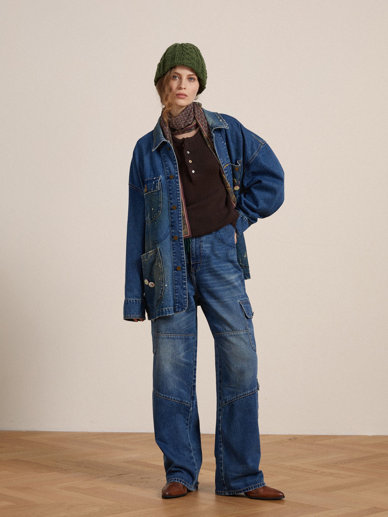 A woman wearing a denim jacket and Siwa Cargo Paneled Jeans, a brown sweater, green beanie, and brown shoes, stands confidently in a room with a wood floor.