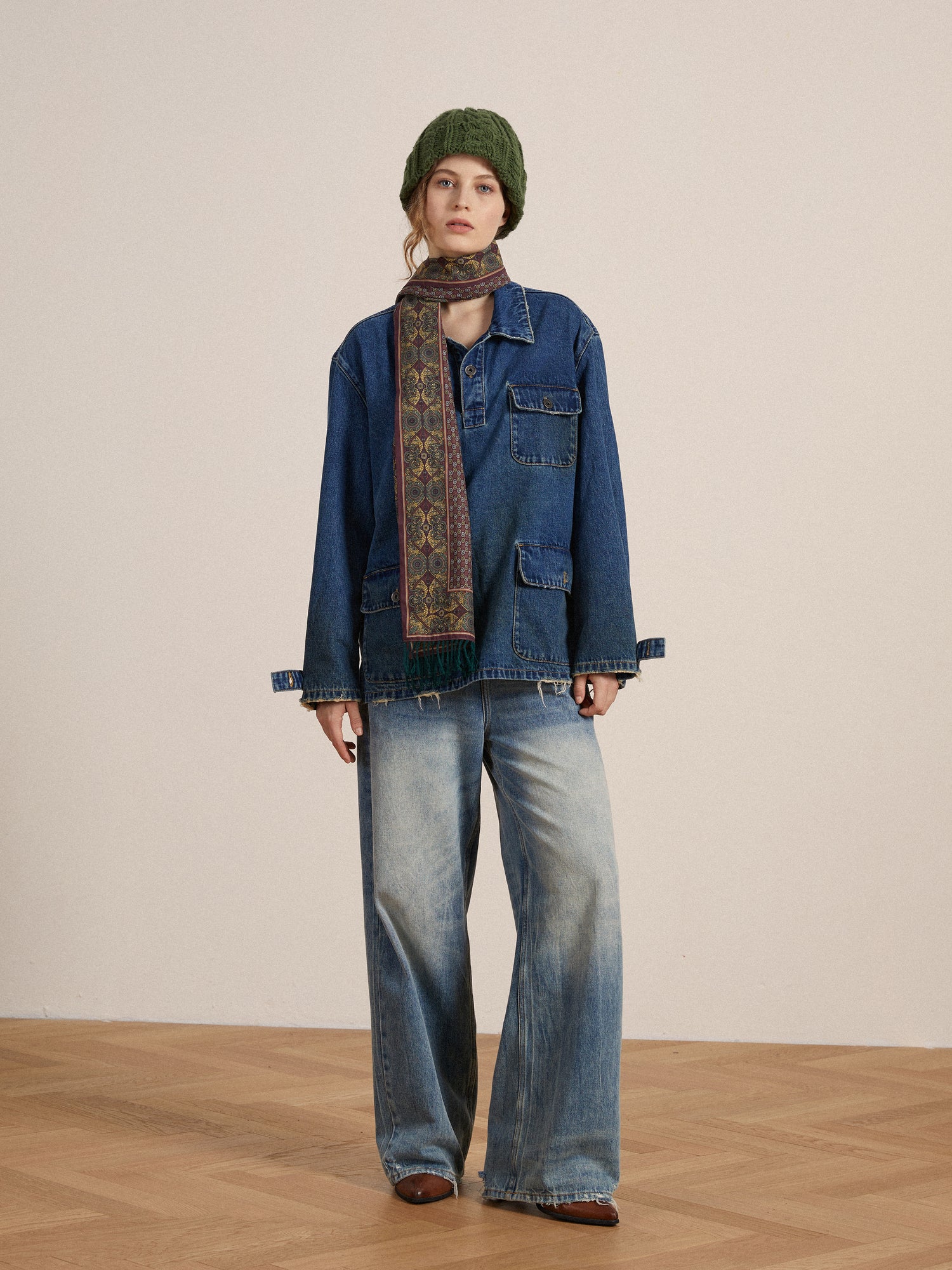 A woman stands in a studio, wearing a Found Azin Denim Pullover Work Shirt, wide-leg jeans, a patterned scarf, and a green beanie, looking at the camera with a neutral expression.