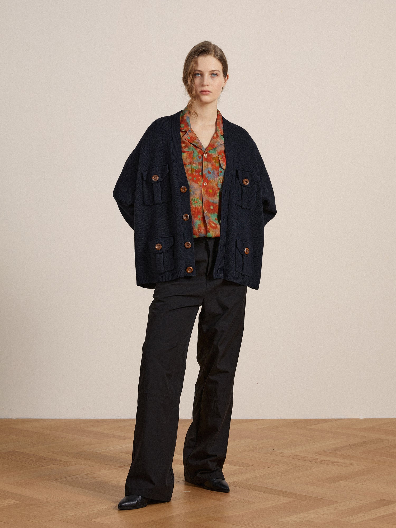 A woman in a stylish oversized navy blue cardigan, colorful blouse, and Found Tencel Pleated Pants, standing in a neutral-toned room.