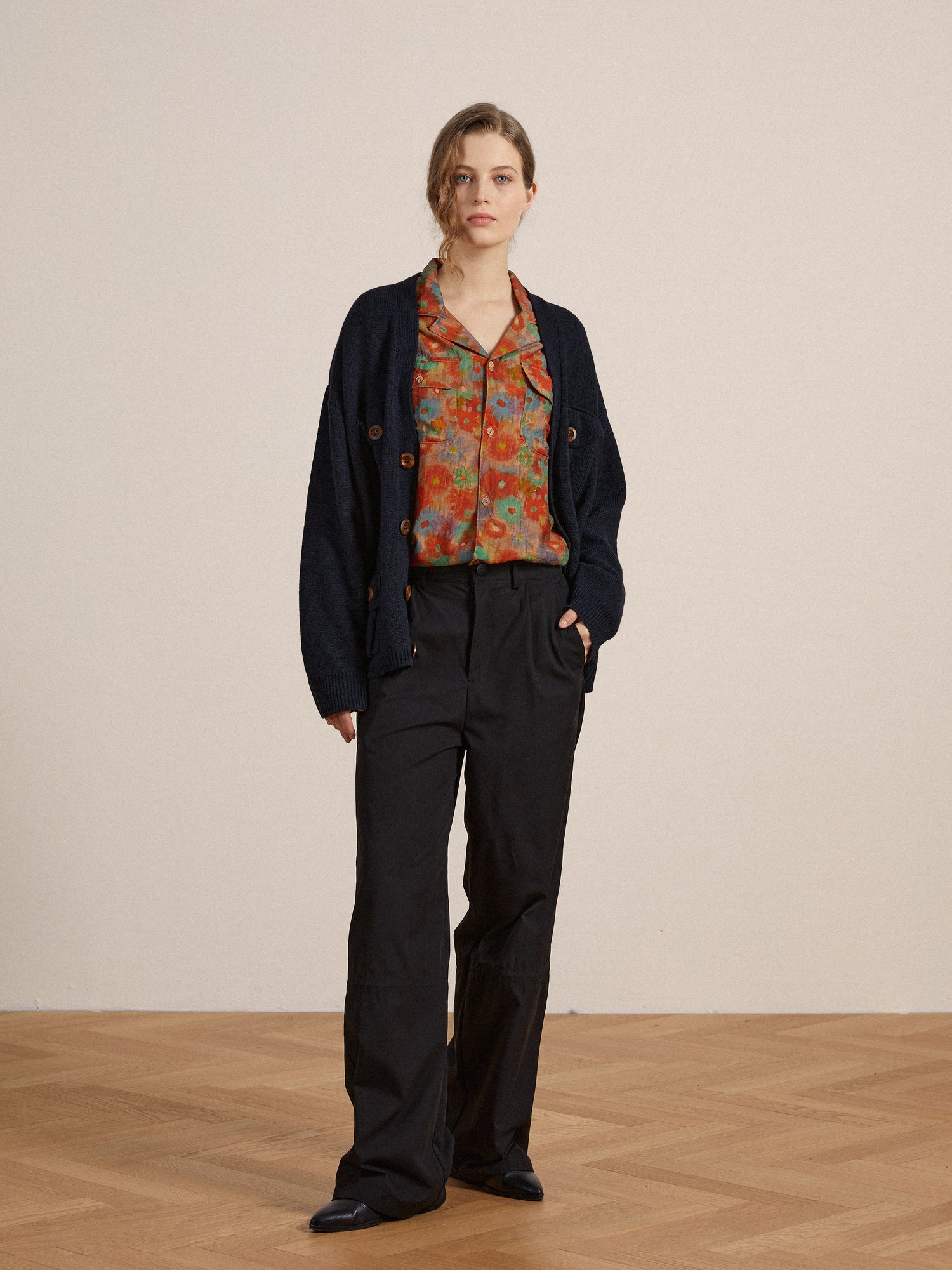 A woman stands in a neutral-toned room wearing a navy cardigan, colorful blouse, and Found Tencel Pleated Pants, looking directly at the camera.