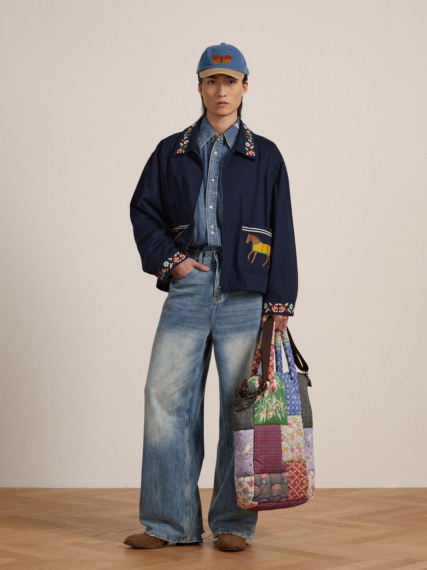 Man in eclectic outfit featuring a Found Horse Equine Work Jacket with detailed embroidery, wide-leg jeans, a baseball cap, and a colorful patchwork tote bag, standing against a plain backdrop.