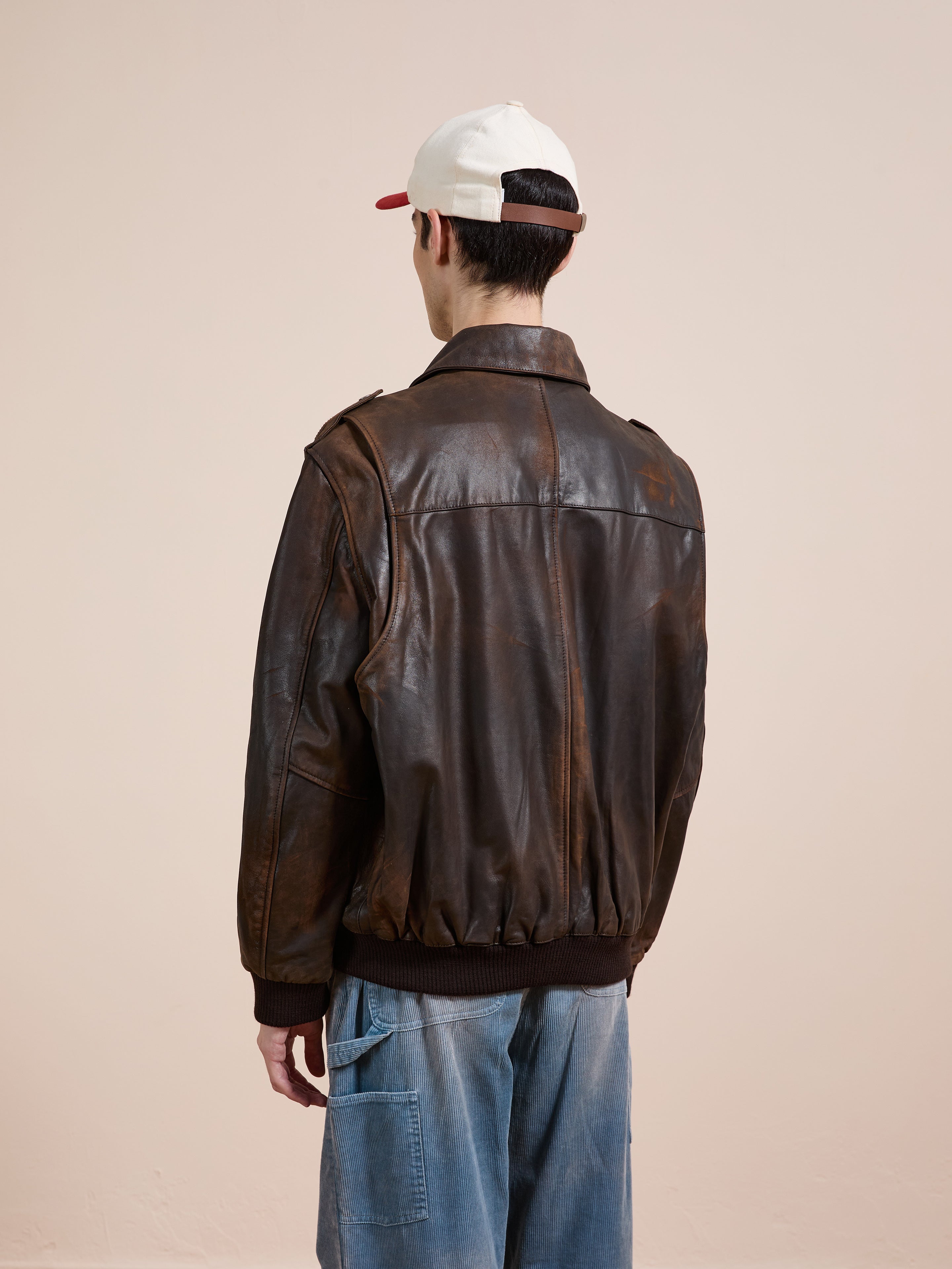 A person wearing blue jeans, a white baseball cap, and the FOUND Distressed Leather Flight Jacket stands facing away from the camera against a plain beige background.
