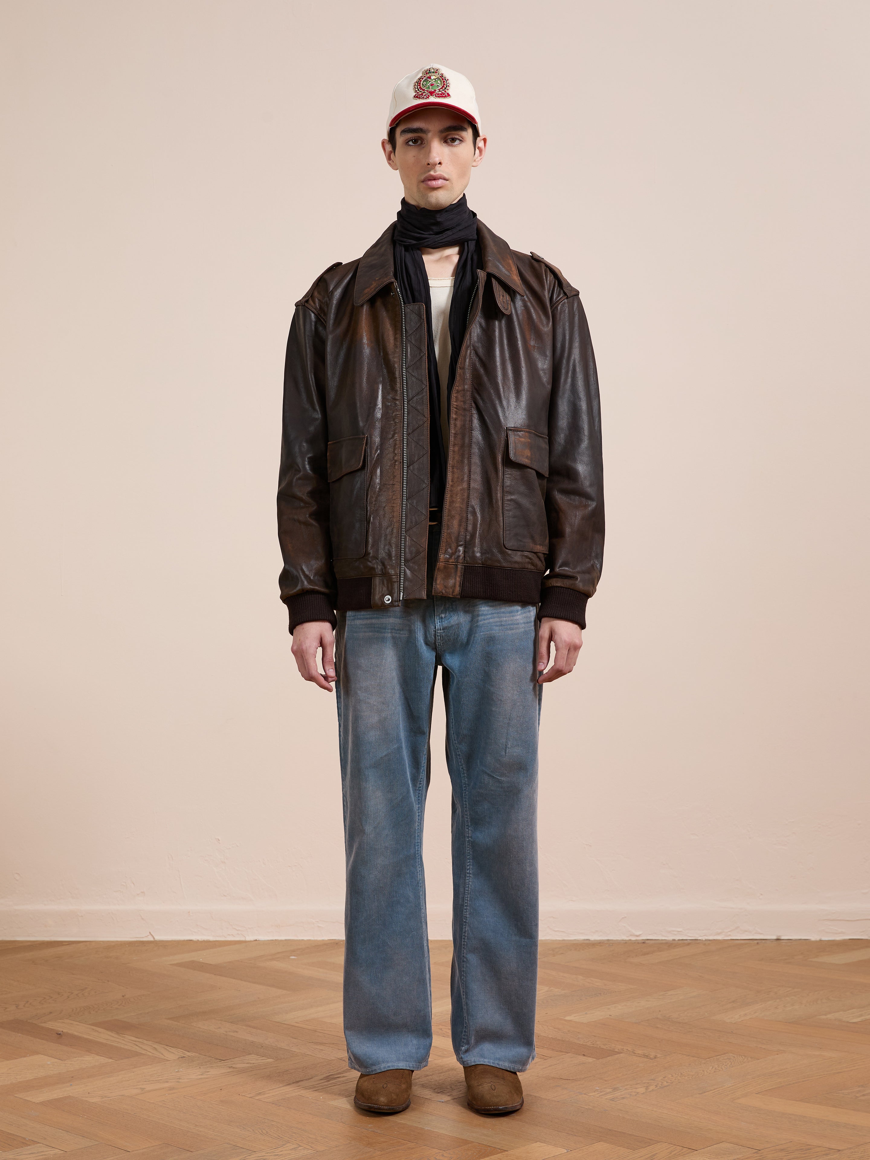 A person stands indoors wearing a white cap, black scarf, FOUND Distressed Leather Flight Jacket, light blue jeans, and brown shoes on a wooden floor with a plain light wall behind.
