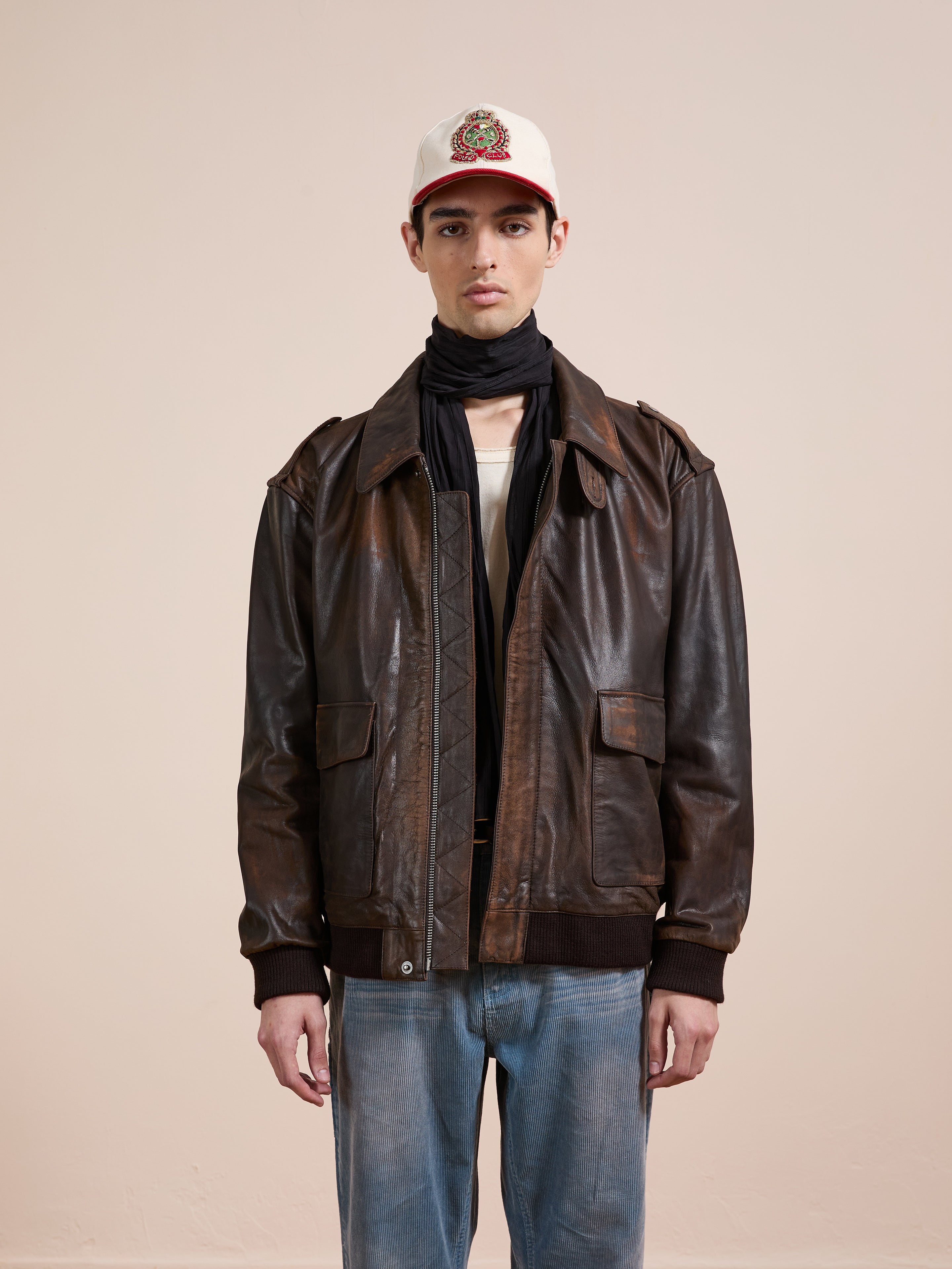 A person stands against a plain background wearing the FOUND Distressed Leather Flight Jacket, light blue jeans, a white logo cap, and a black scarf.