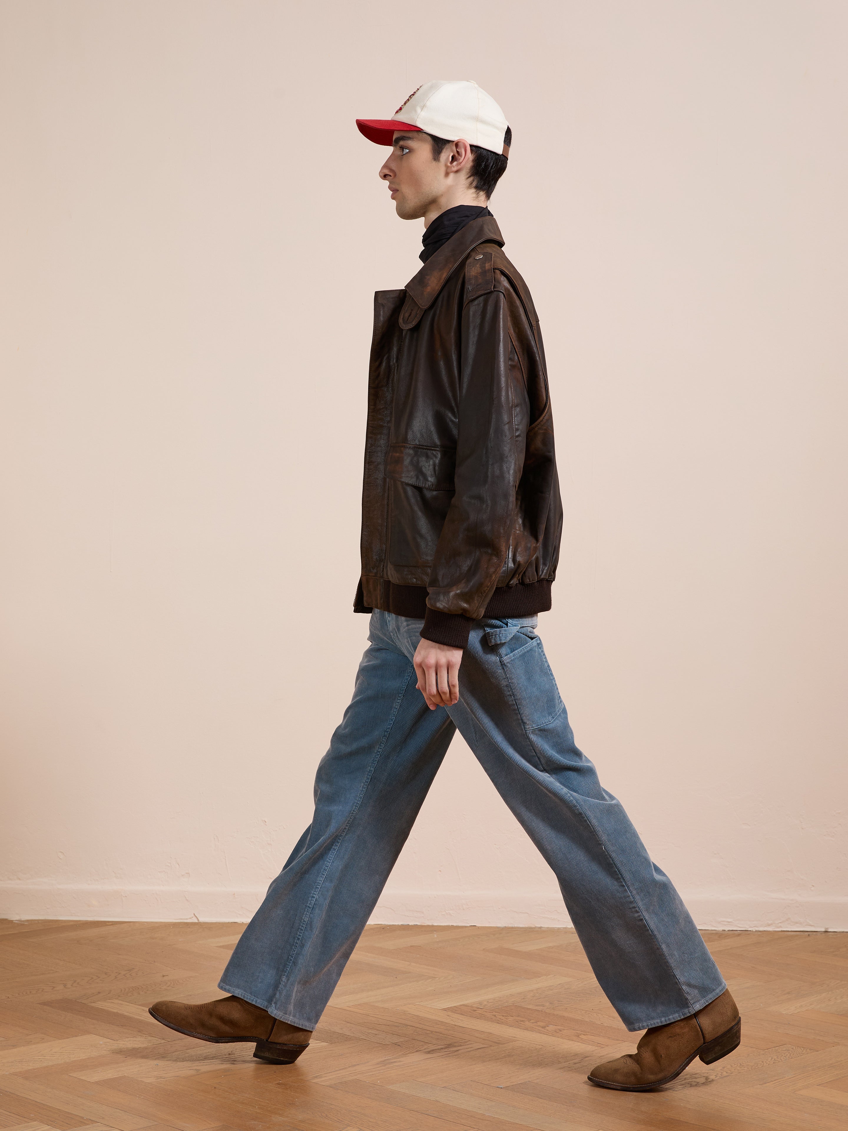 Wearing a FOUND Distressed Leather Flight Jacket, blue jeans, brown boots, and a white and red cap, a person walks indoors against a plain beige wall.
