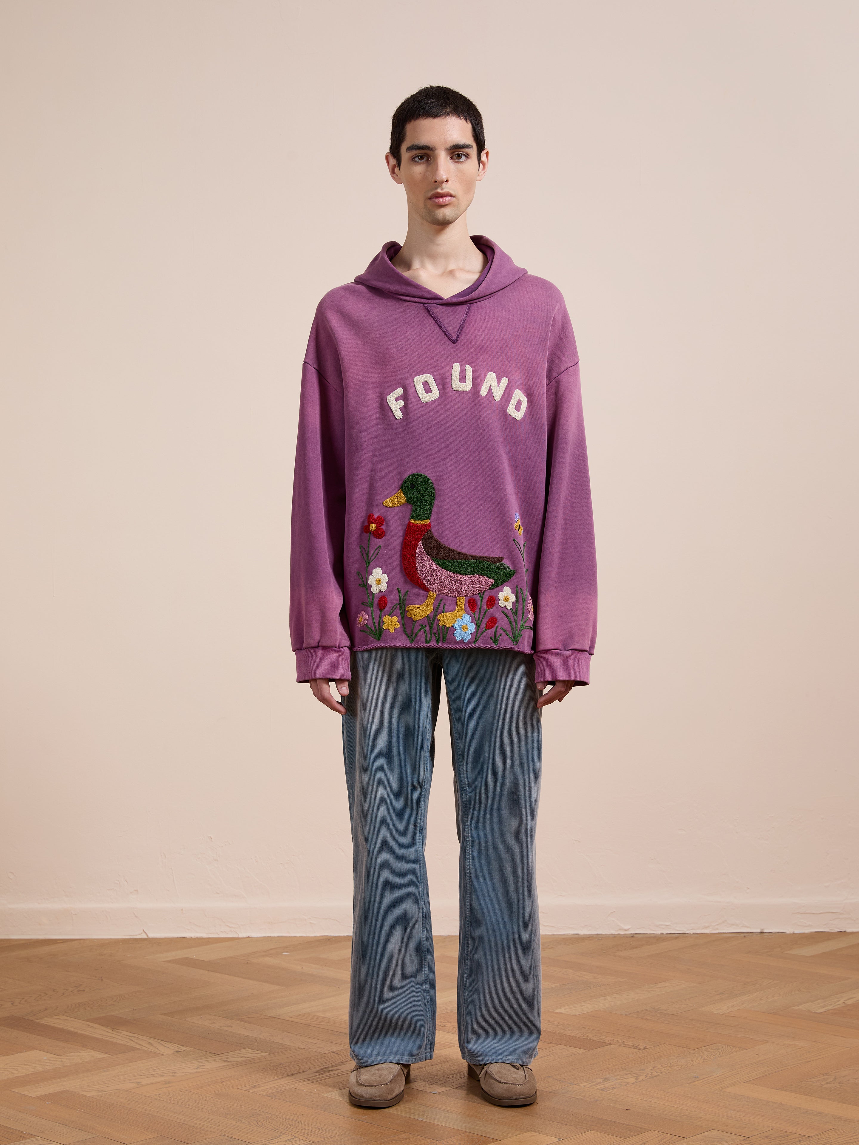 A person stands indoors against a plain wall and parquet floor, wearing the FOUND Embroidered Wildlife Hoodie in purple with duck embroidery, paired with blue jeans and beige shoes.