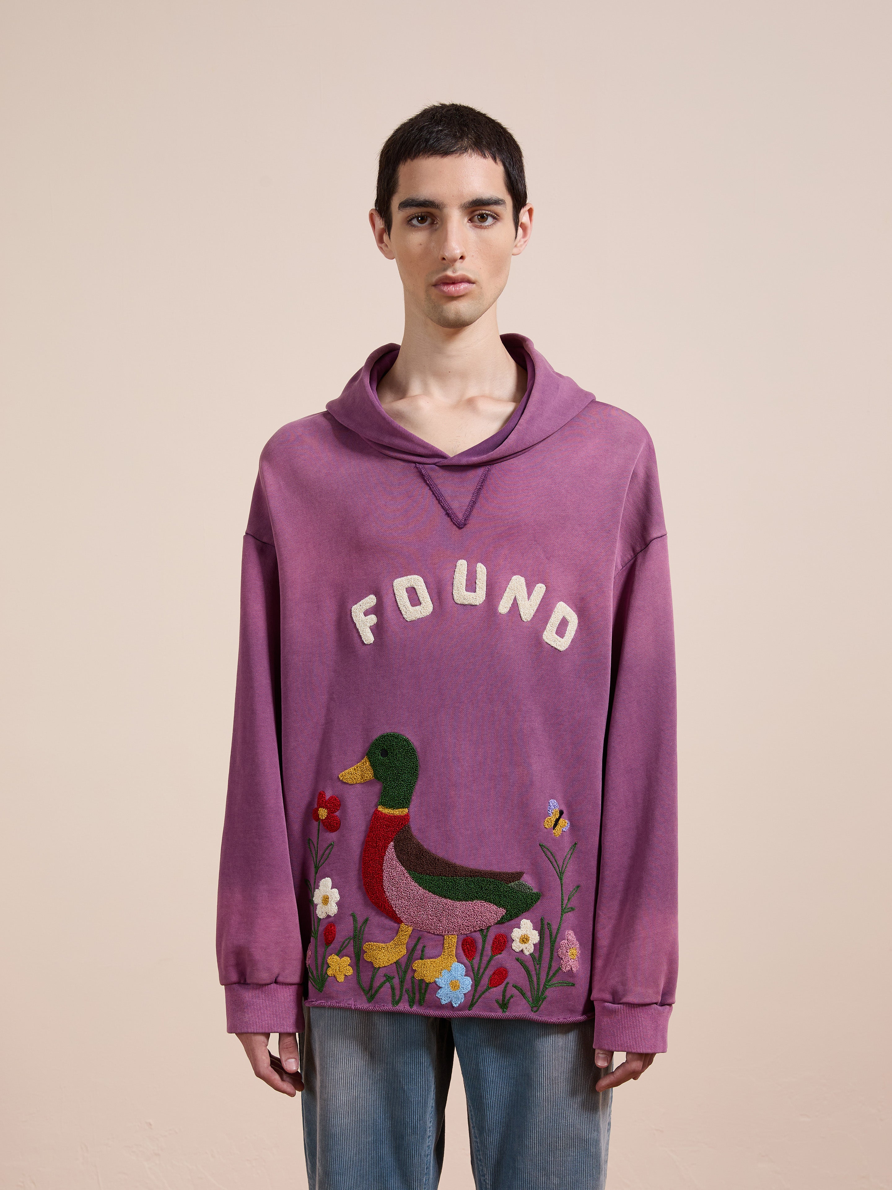 A young person with short dark hair wears the FOUND Embroidered Wildlife Hoodie, featuring an embroidered duck and flowers, standing against a plain beige background.