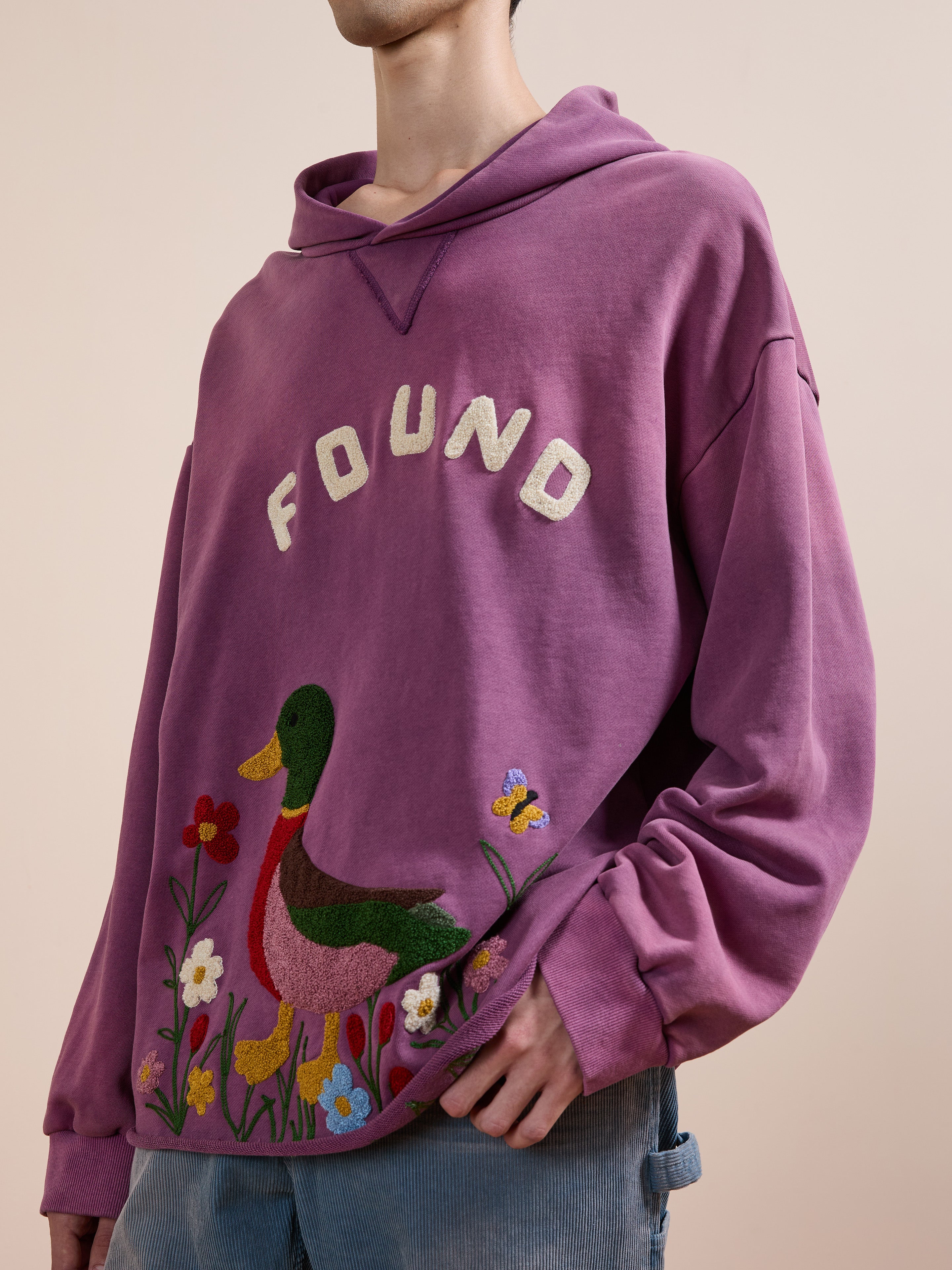 A person wearing the FOUND Embroidered Wildlife Hoodie, featuring colorful duck, flower, and butterfly designs on purple French-terry cotton. Only the lower half of their face is visible.