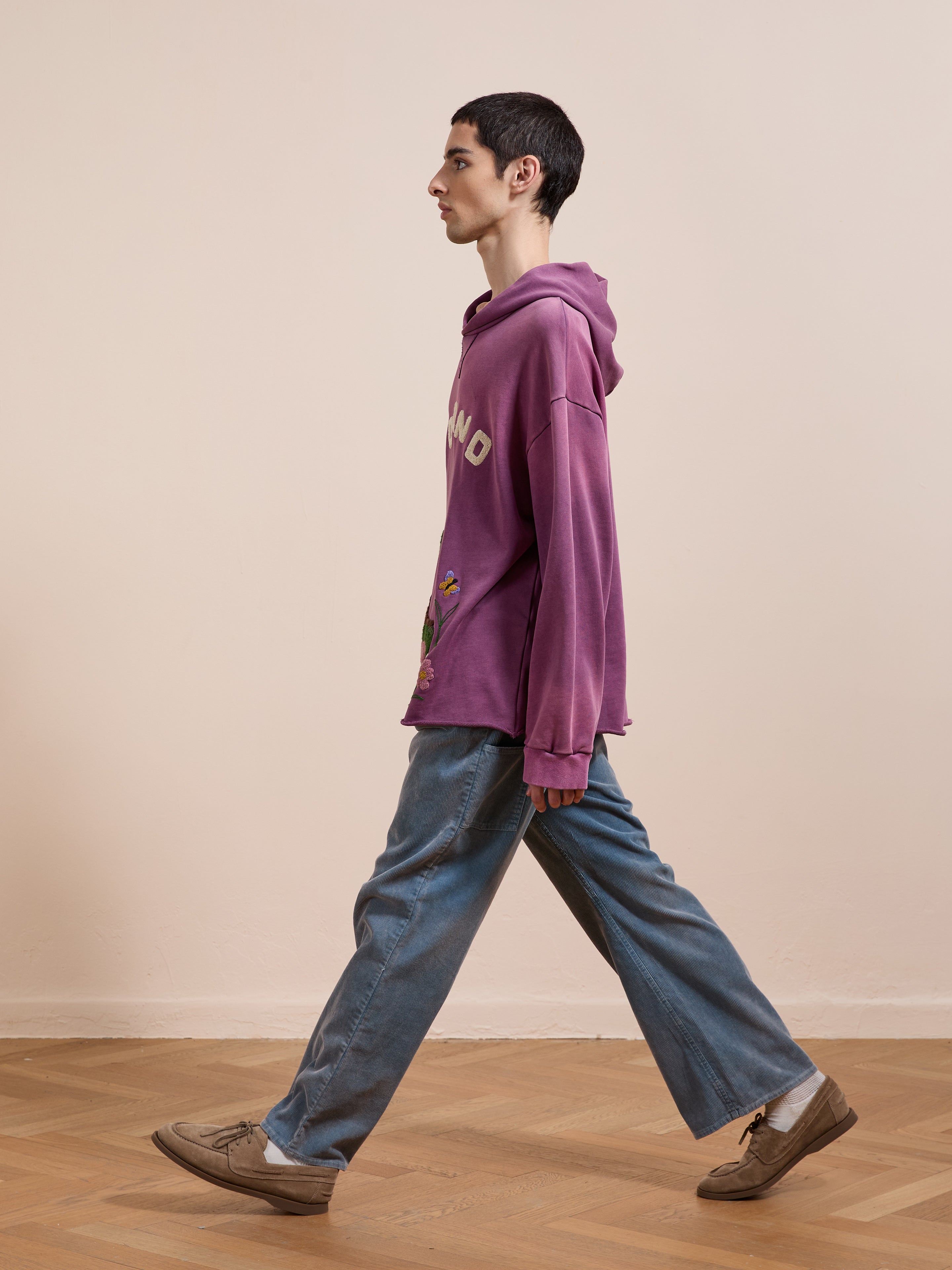 A young person in the FOUND Embroidered Wildlife Hoodie and blue jeans walks indoors across a wooden floor, facing left against a plain beige wall.