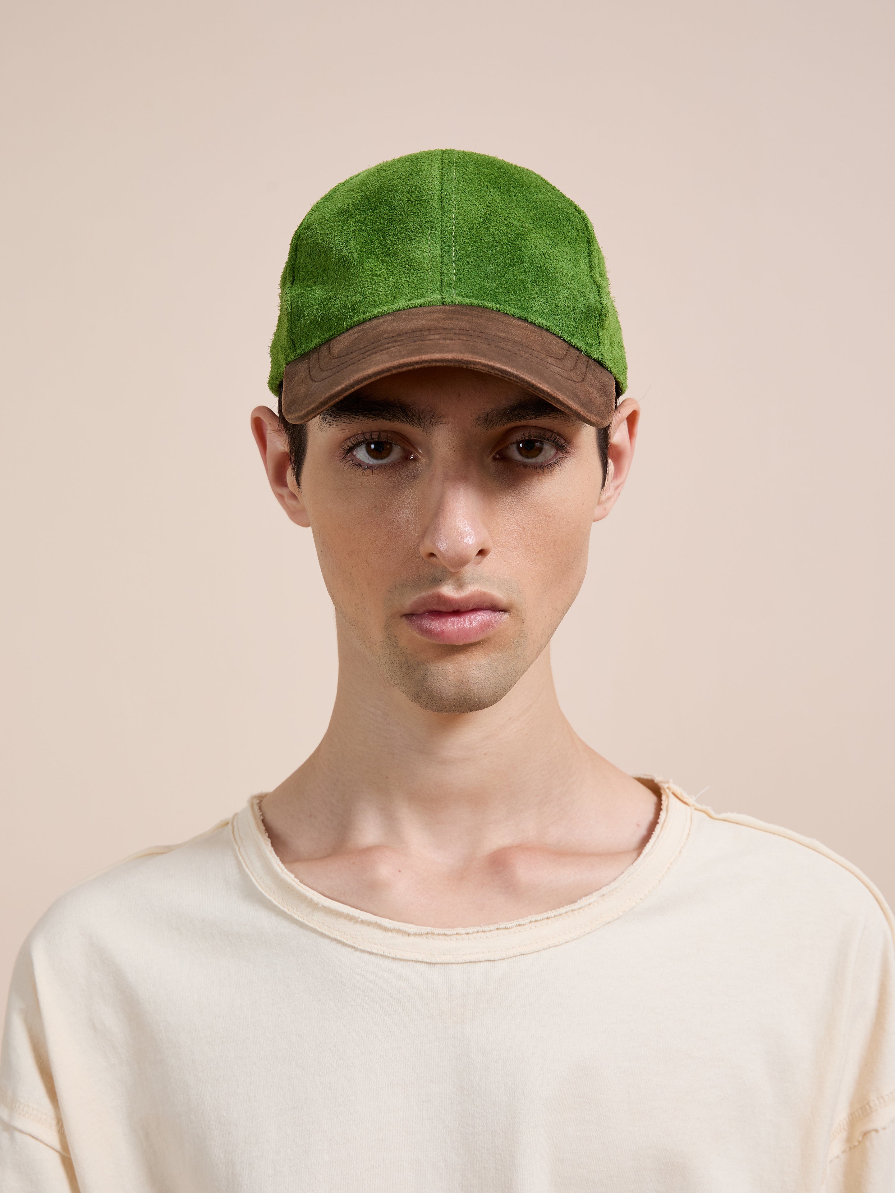 A person wearing the Found Suede Leather Contrast Distressed Cap, featuring green suede and a distressed brown leather brim, paired with a cream shirt, faces forward against a plain beige background.