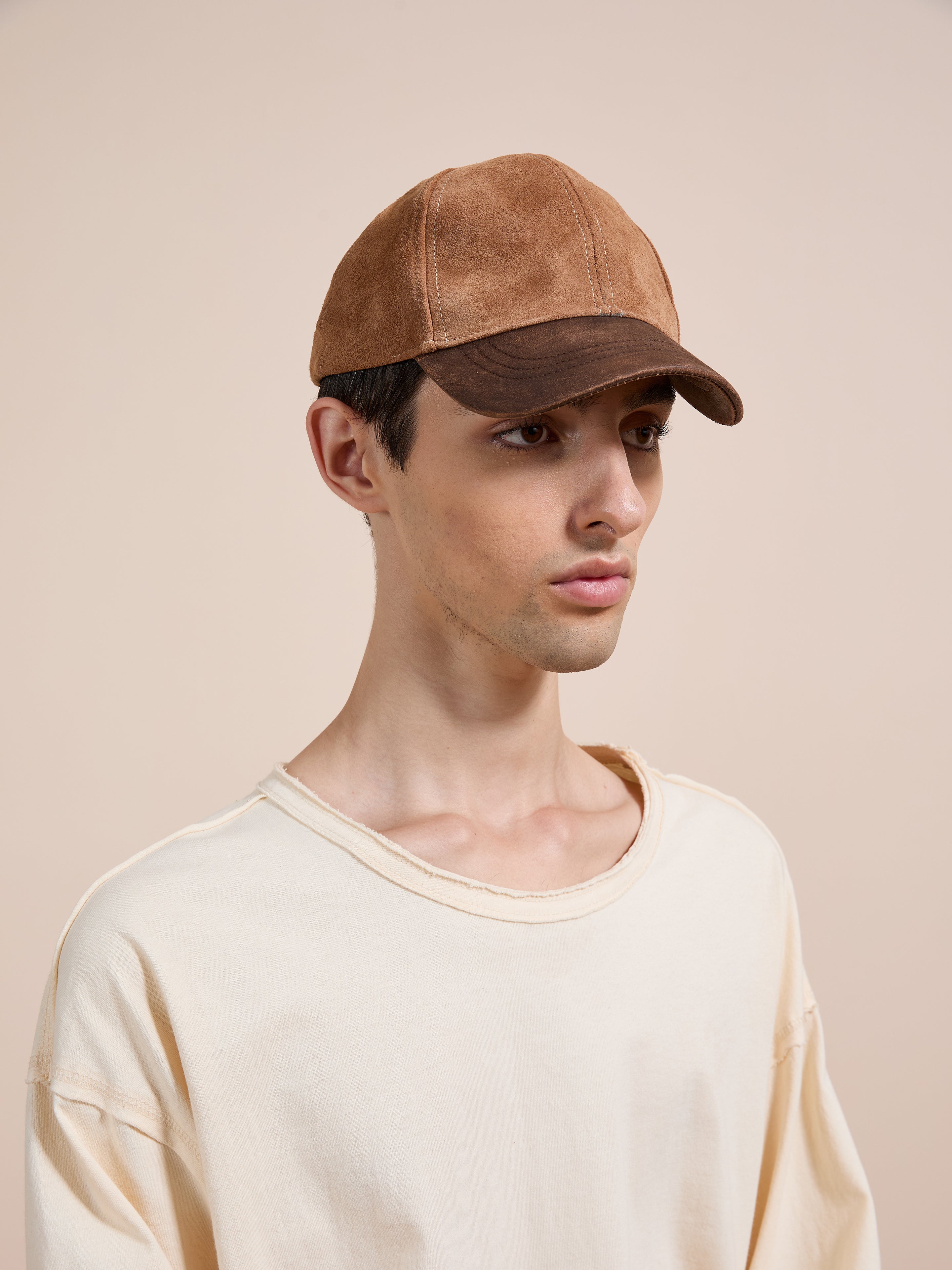 A young person wears the Found Suede Leather Contrast Distressed Cap and an off-white oversized shirt, standing against a plain beige background.