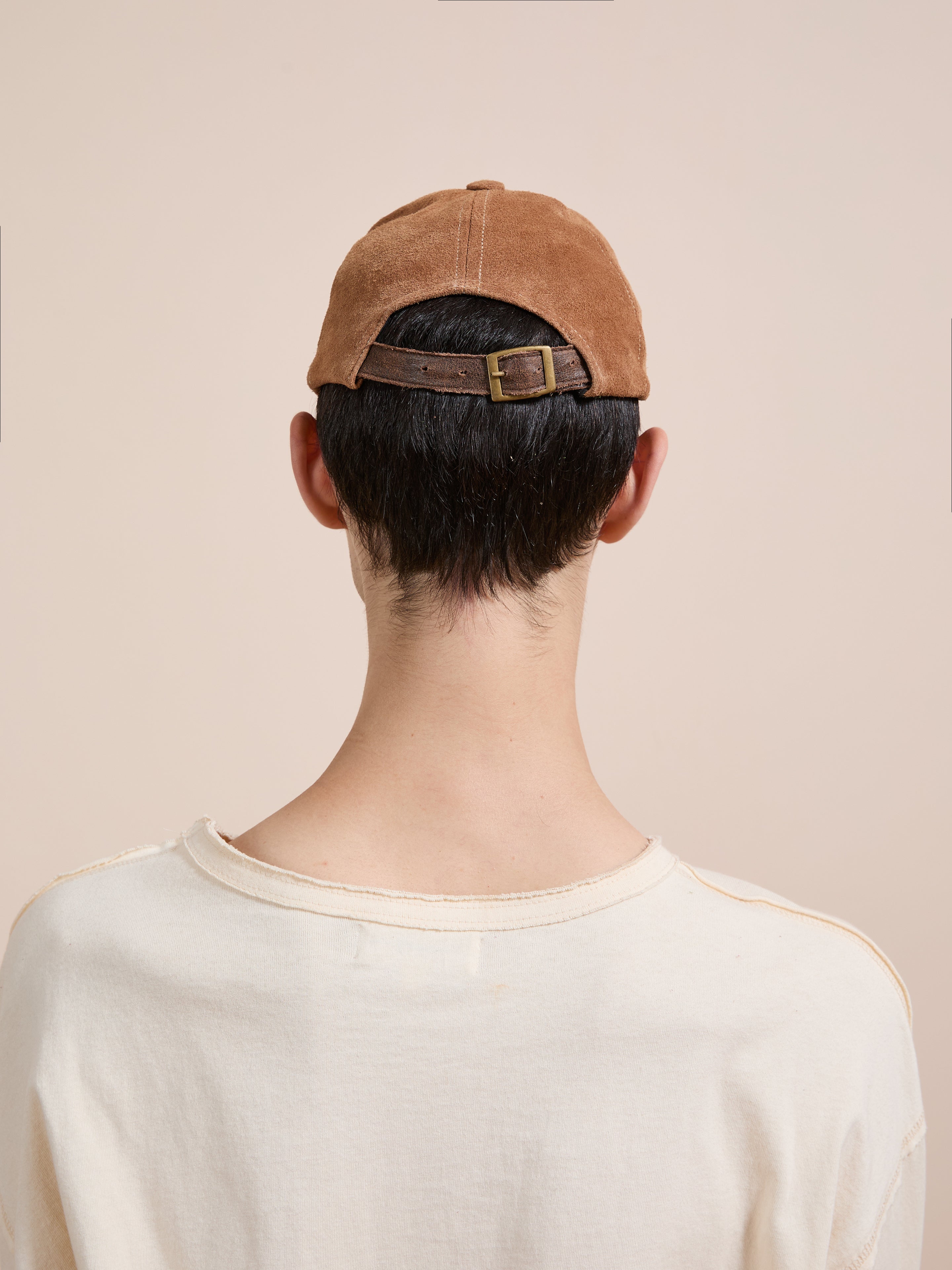 A person with short dark hair wears the Found Suede Leather Contrast Distressed Cap, featuring an adjustable leather strap, paired with a beige shirt, shown from behind against a plain light background.