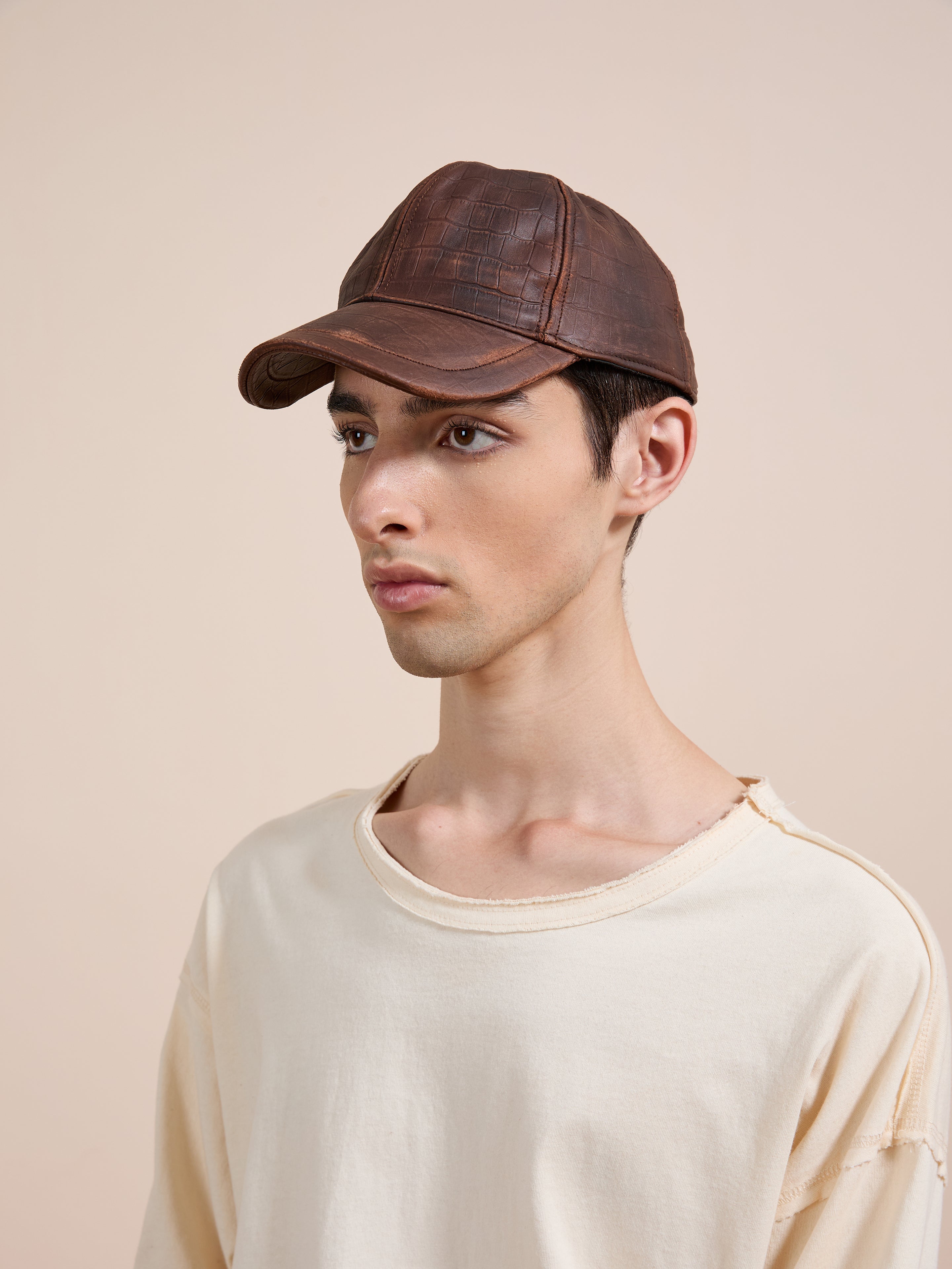 A young person wears the Found Croc Embossed Distressed Leather Cap with an off-white, lightly distressed tee against a plain beige background.