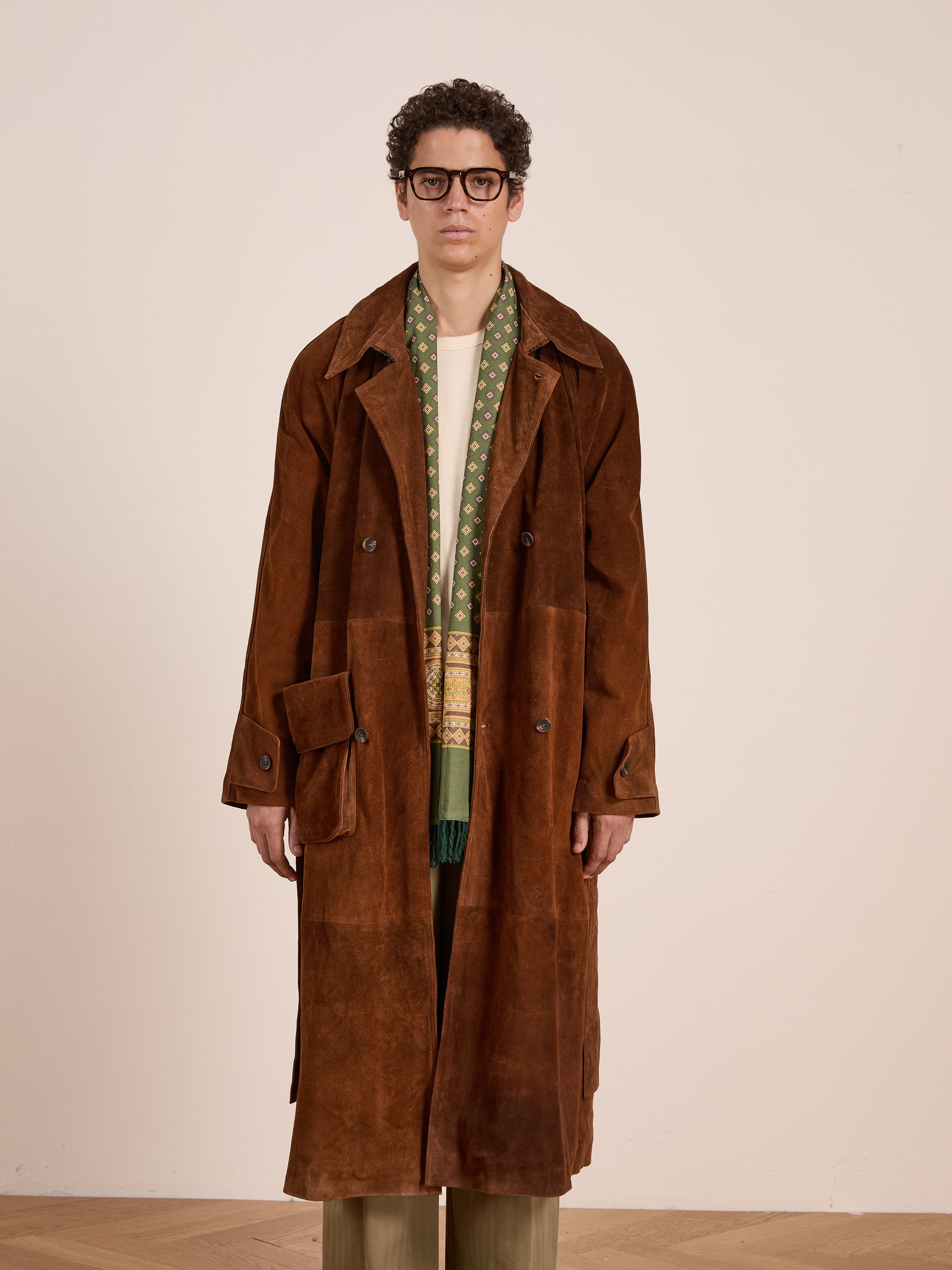 A person wearing glasses and a FOUND Suede Leather Trench Coat in brown, paired with a green patterned scarf, light shirt, and olive trousers, stands against a plain light-colored background.