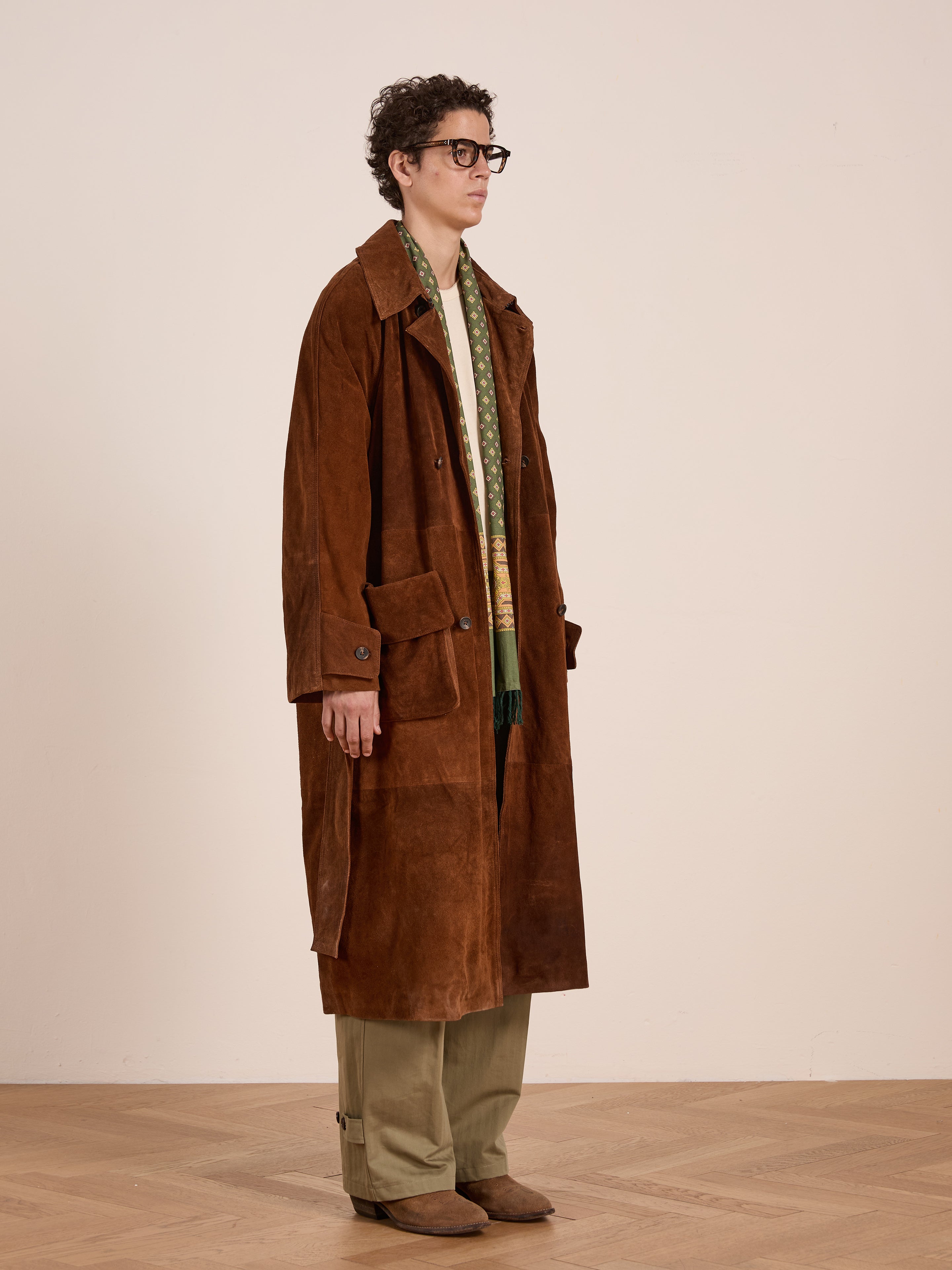 A person in glasses wears the FOUND Suede Leather Trench Coat, wide-leg khaki pants, brown shoes, and a green scarf while standing indoors against a plain wall.