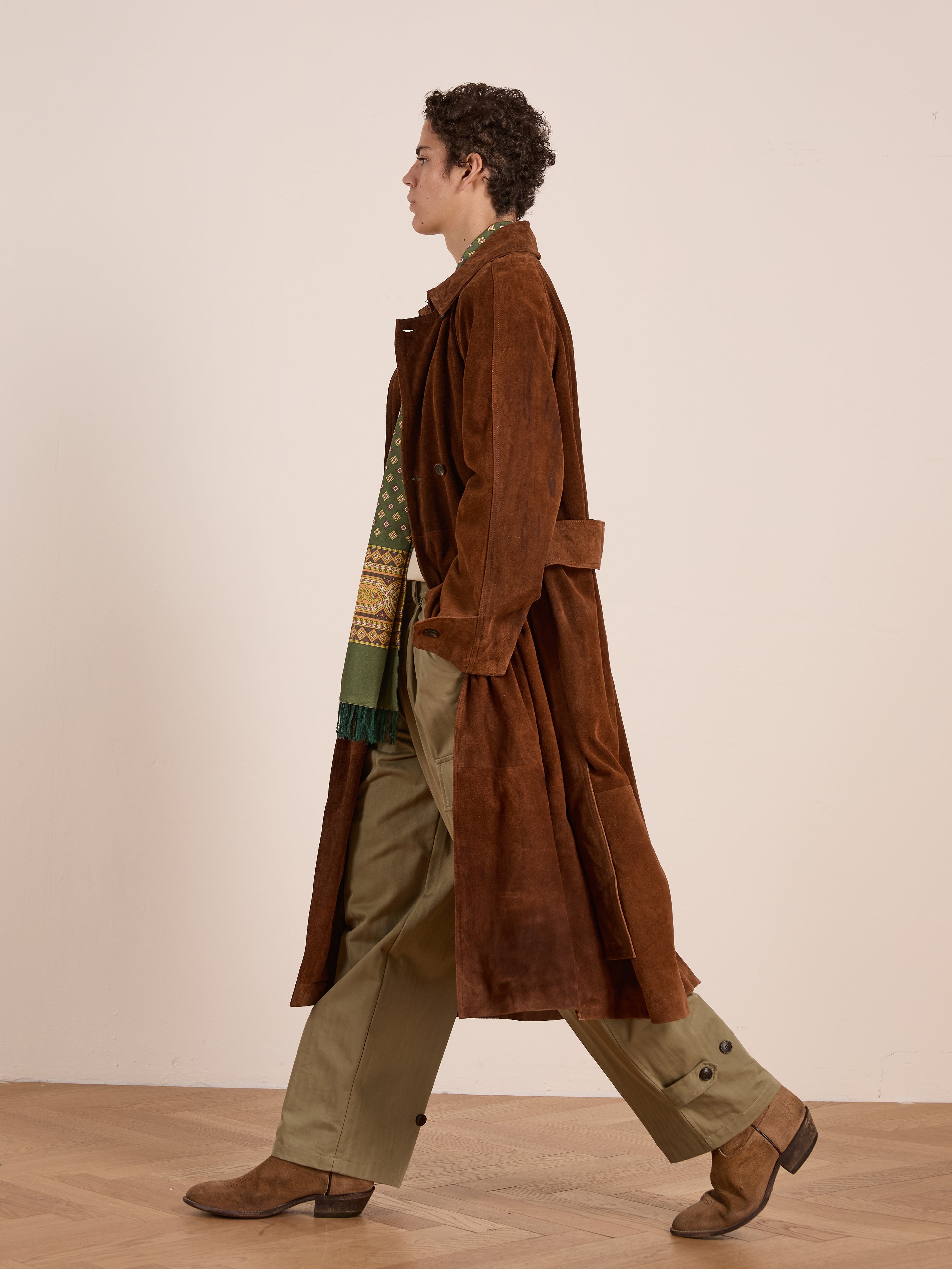 A person walks indoors wearing the FOUND Suede Leather Trench Coat, olive green pants, tan boots, and a green and yellow patterned scarf.