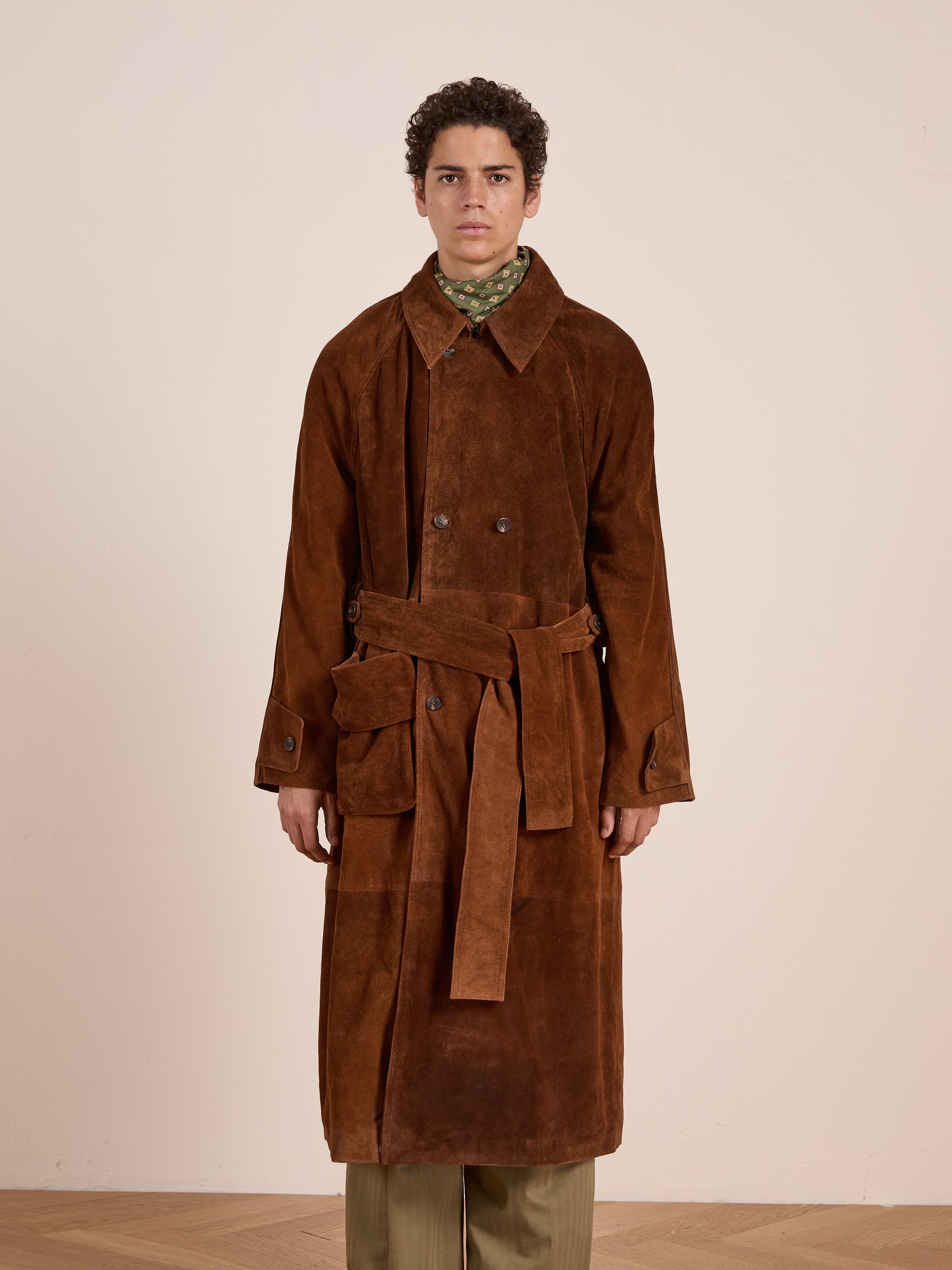 A person stands indoors wearing the FOUND Suede Leather Trench Coat—an ethically crafted, unisex brown suede coat with a belt and pockets—paired with beige pants.
