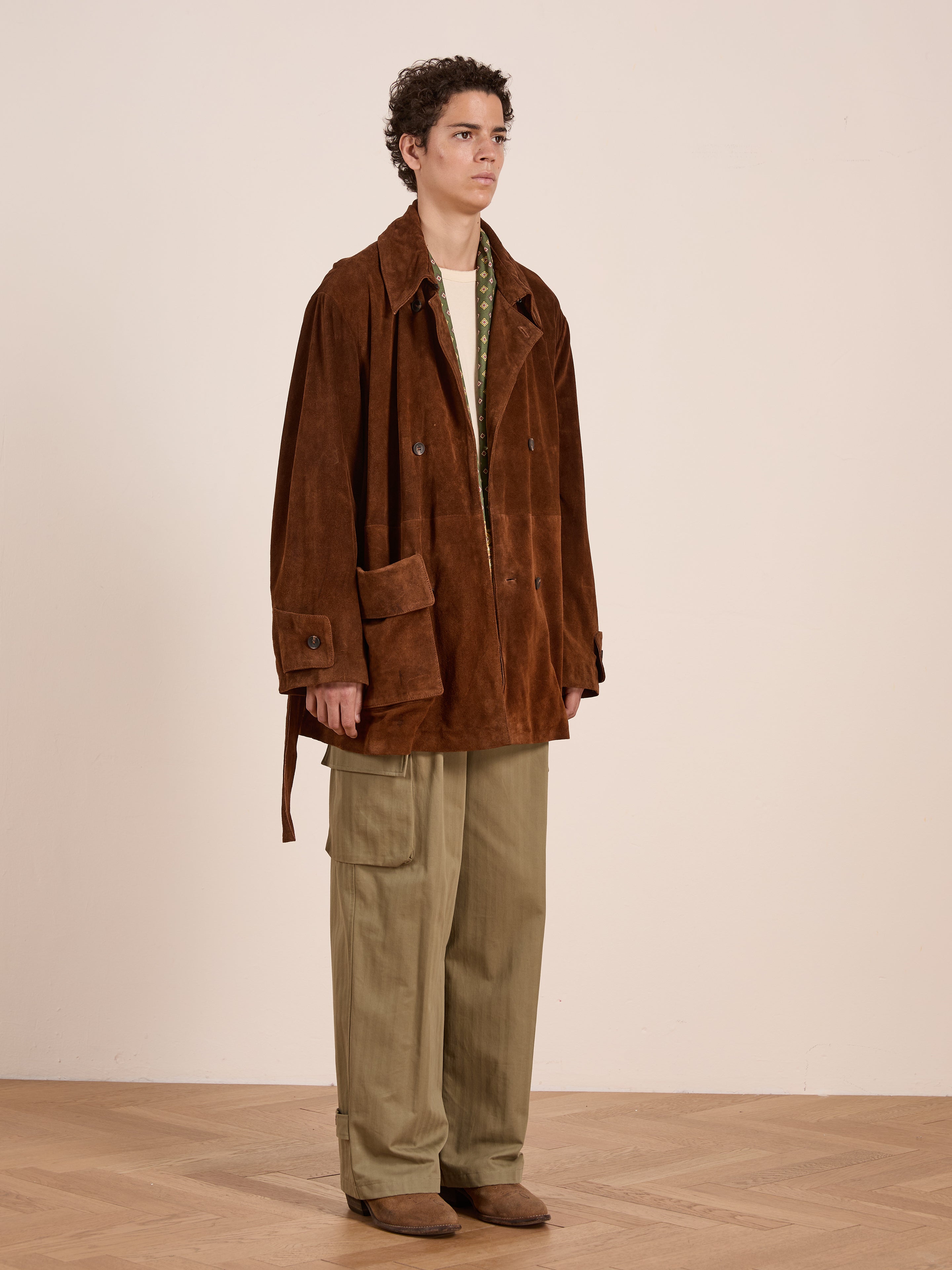 Indoors against a plain light wall, a person wears the FOUND Double Breasted Suede Leather Jacket with olive green cargo pants, a beige shirt, and brown shoes.