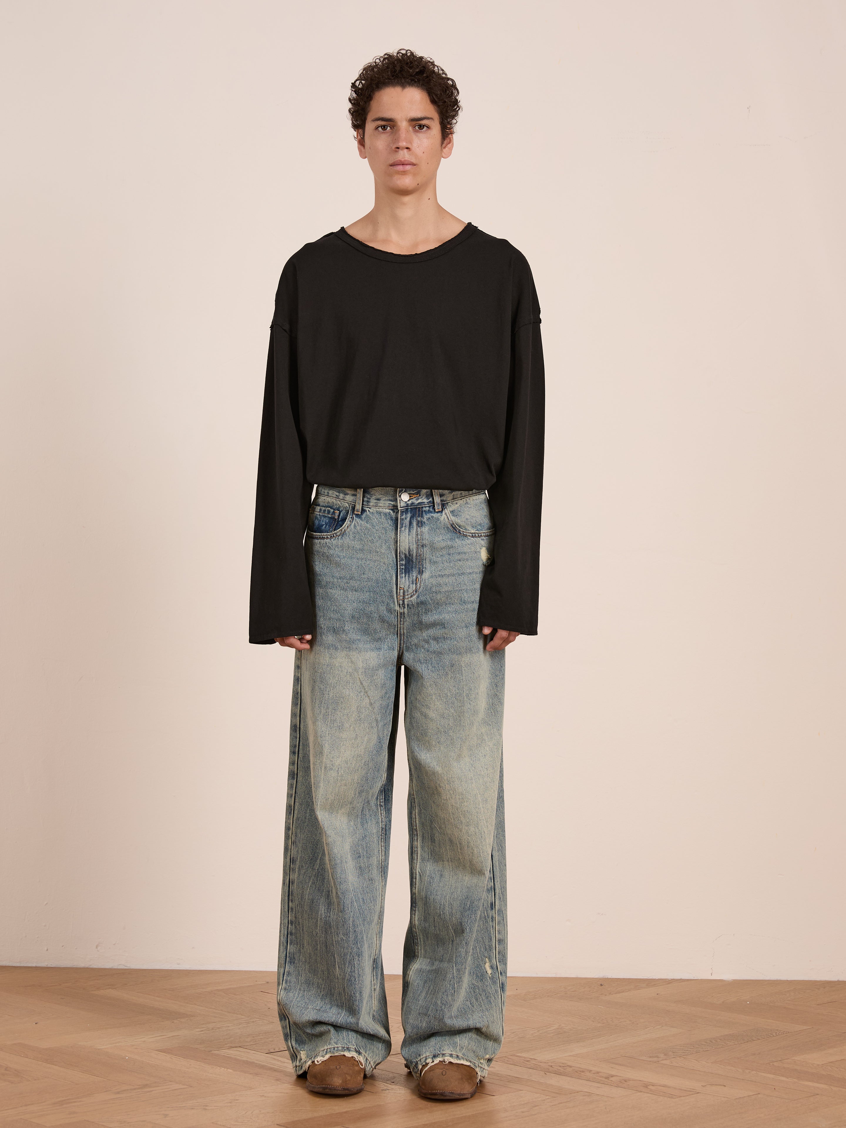 A person stands indoors against a plain wall, wearing a relaxed fit, unisex FOUND Reversed LS Tee in black, paired with baggy blue jeans and brown shoes on a wooden floor.