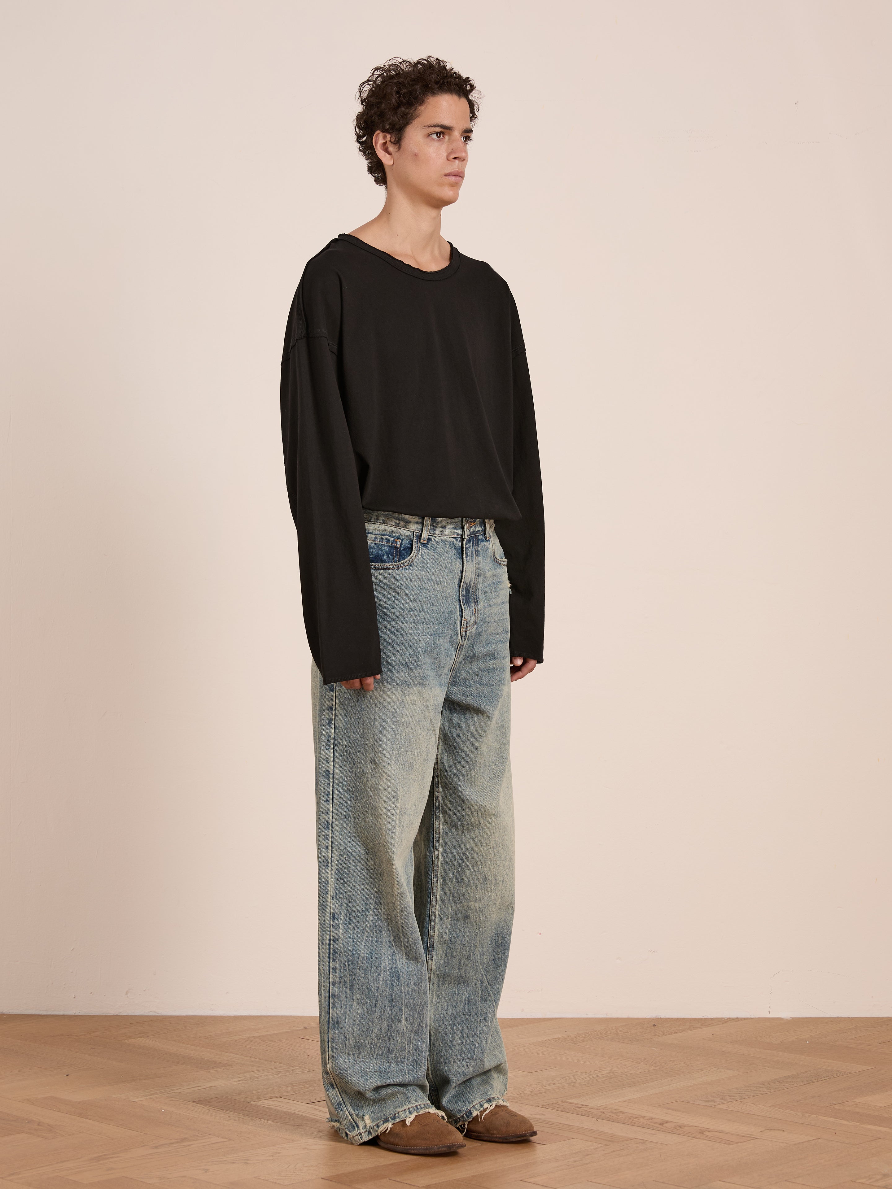 A person stands indoors on a wooden floor against a plain background, wearing a relaxed fit, unisex FOUND Reversed LS Tee with baggy faded blue jeans and brown shoes.