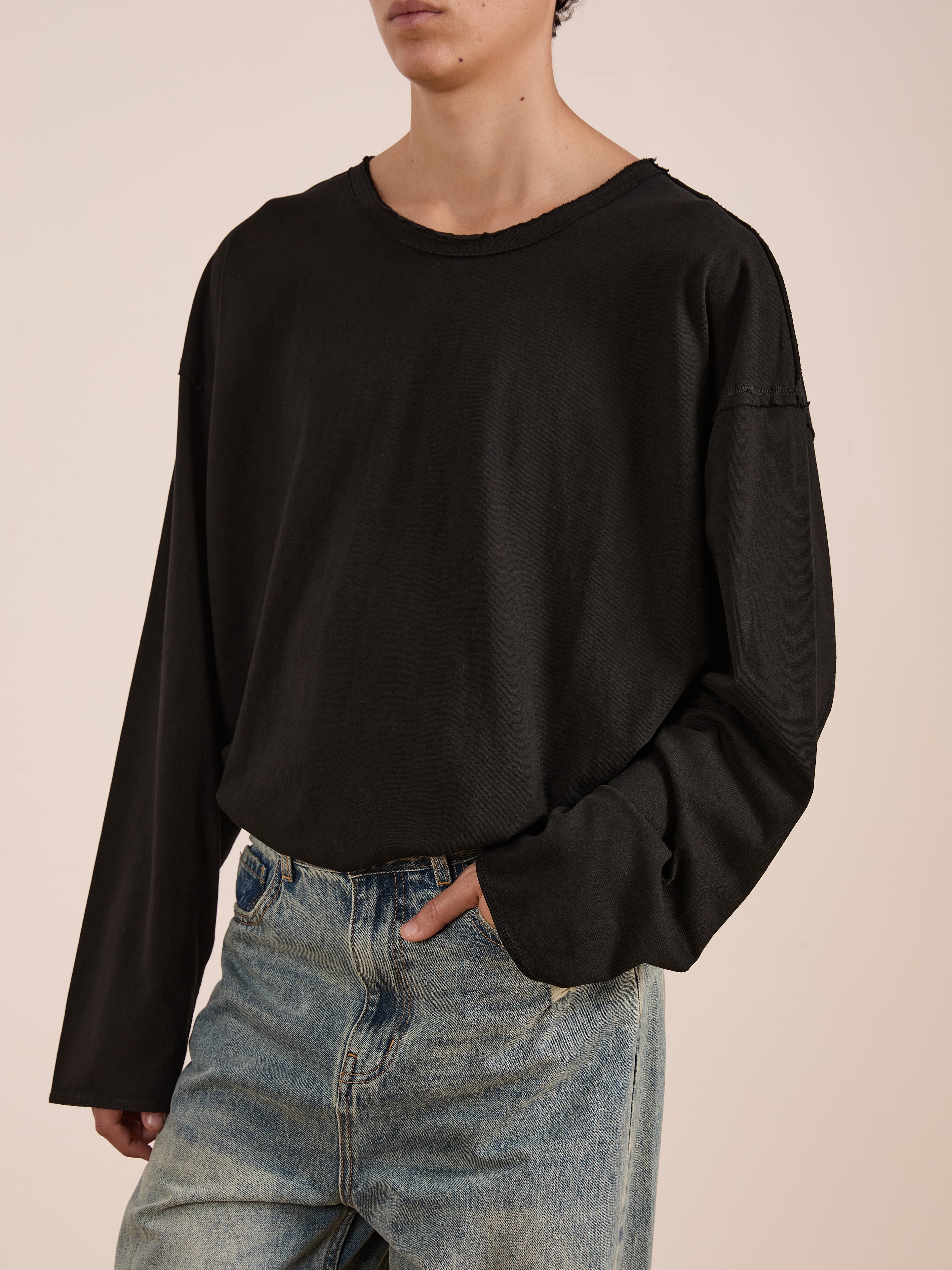 A person stands against a plain background wearing the FOUND Reversed LS Tee in black (relaxed fit, unisex) paired with light blue jeans, one hand in their pocket.