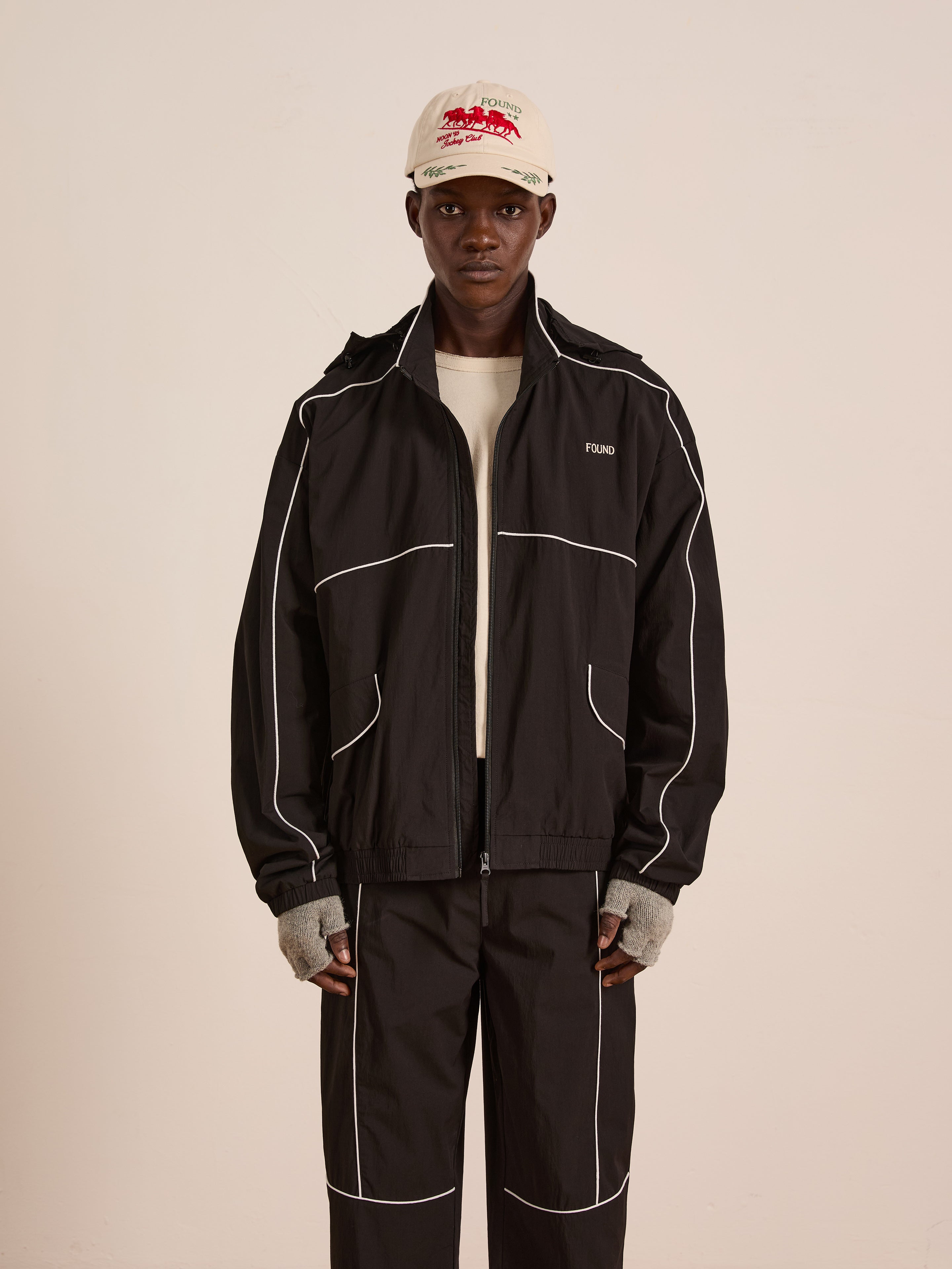 A person stands facing forward in FOUND’s black Arena Stripe Track Jacket and matching pants with white piping, paired with a light shirt, gray fingerless gloves, and a white baseball cap featuring red text.