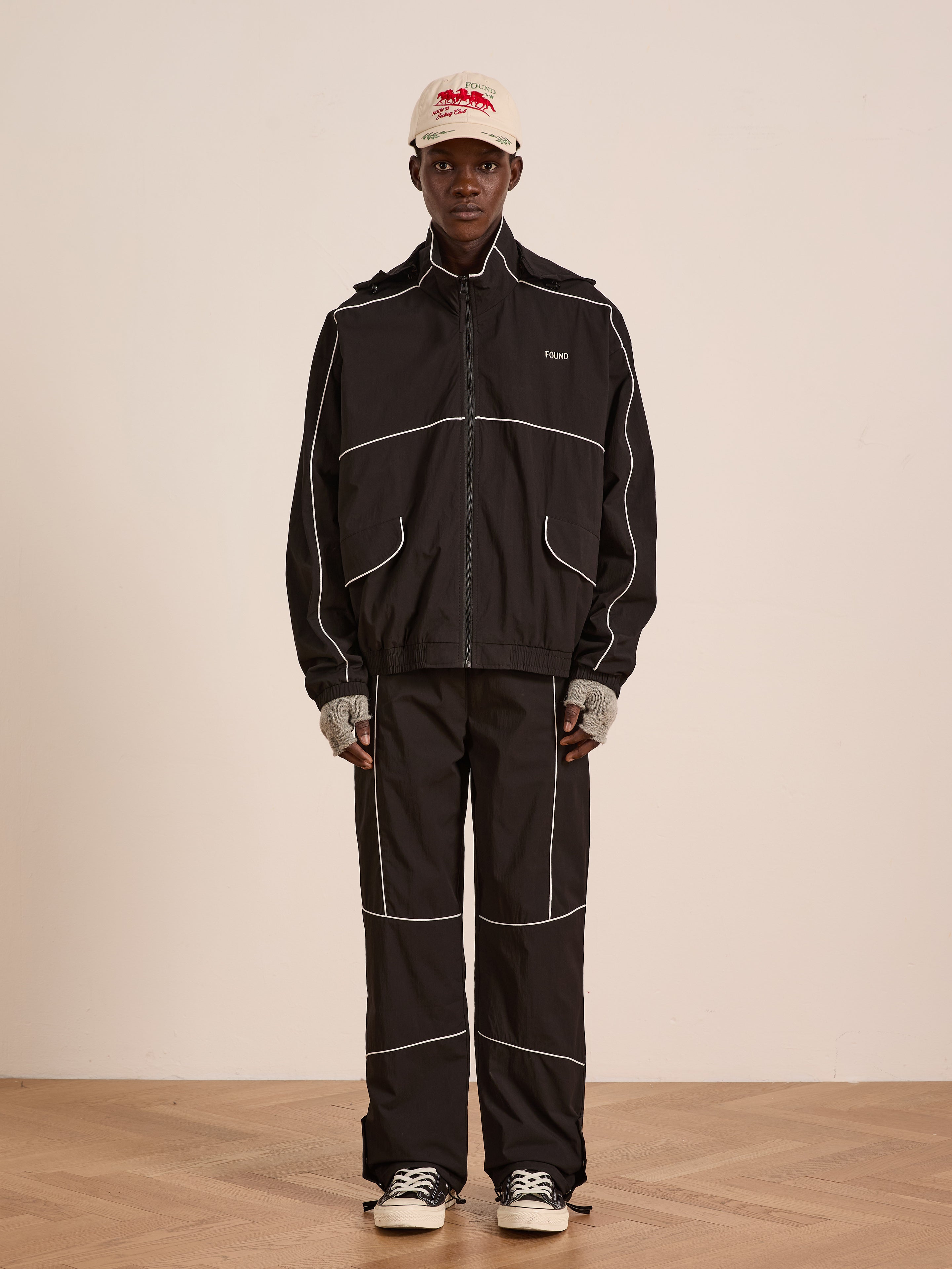 Indoors, a person wears the FOUND Arena Stripe Track Jacket—unisex, black, lightweight technical fabric with white piping—paired with a light cap, gray fingerless gloves, and black-and-white sneakers against a plain background.