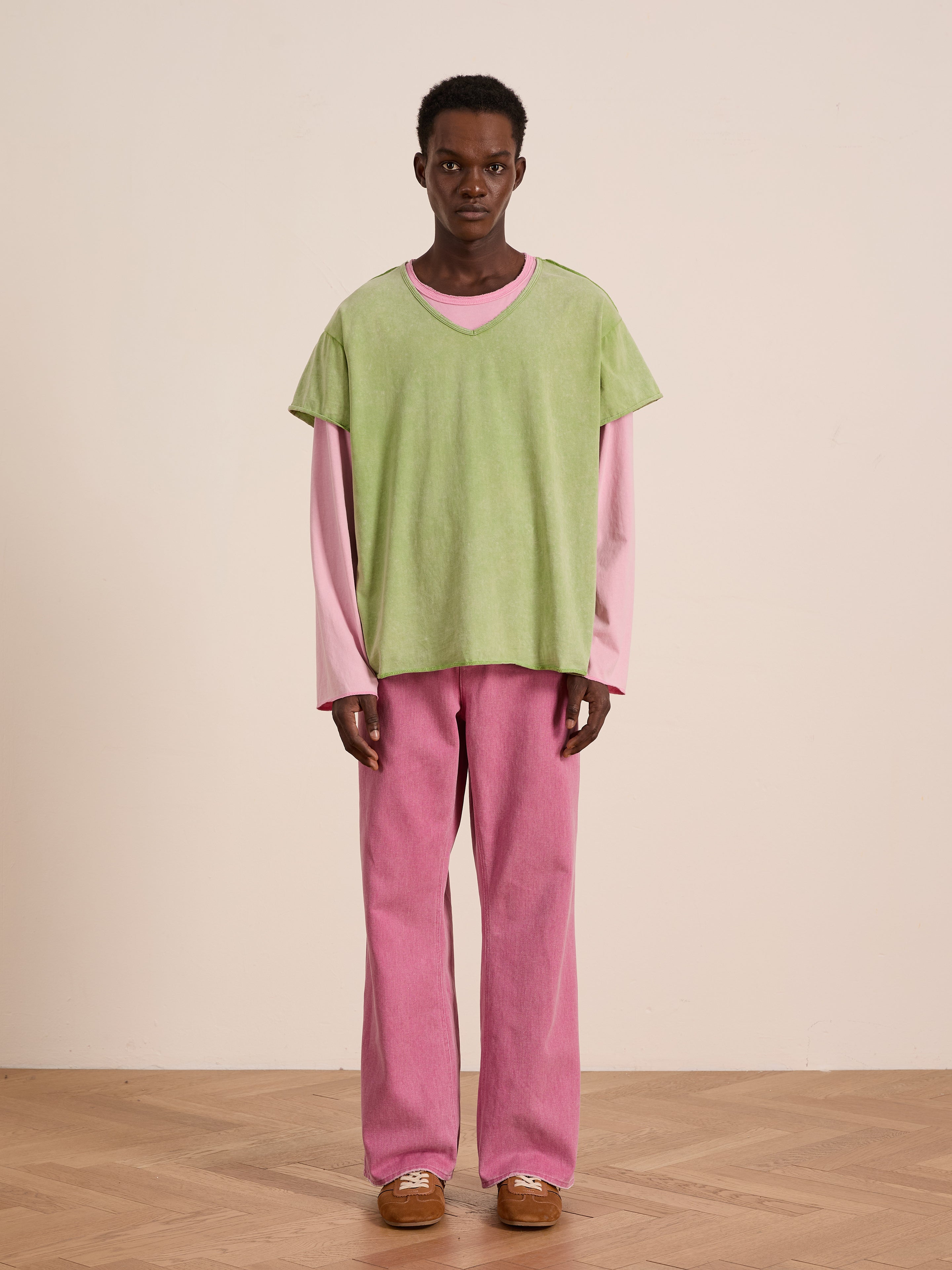 A person stands on a wooden floor wearing FOUND's green Double Layer LS Tee over a pink long sleeve tee, with pink pants and brown shoes, set against a plain beige background.