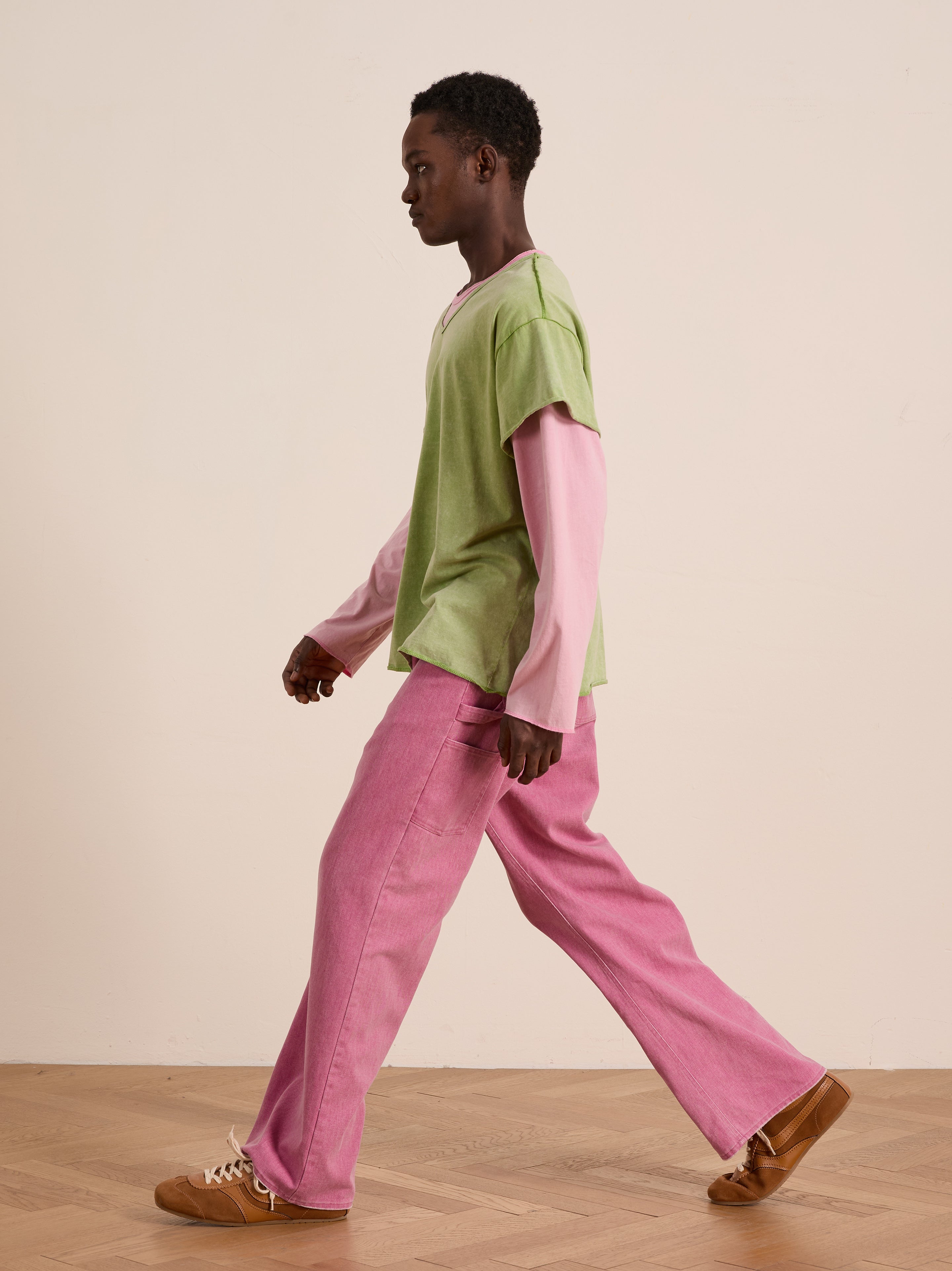 A person walks indoors wearing FOUND’s green Double Layer LS Tee layered over a pink long sleeve tee, paired with pink pants and brown shoes, against a plain background.