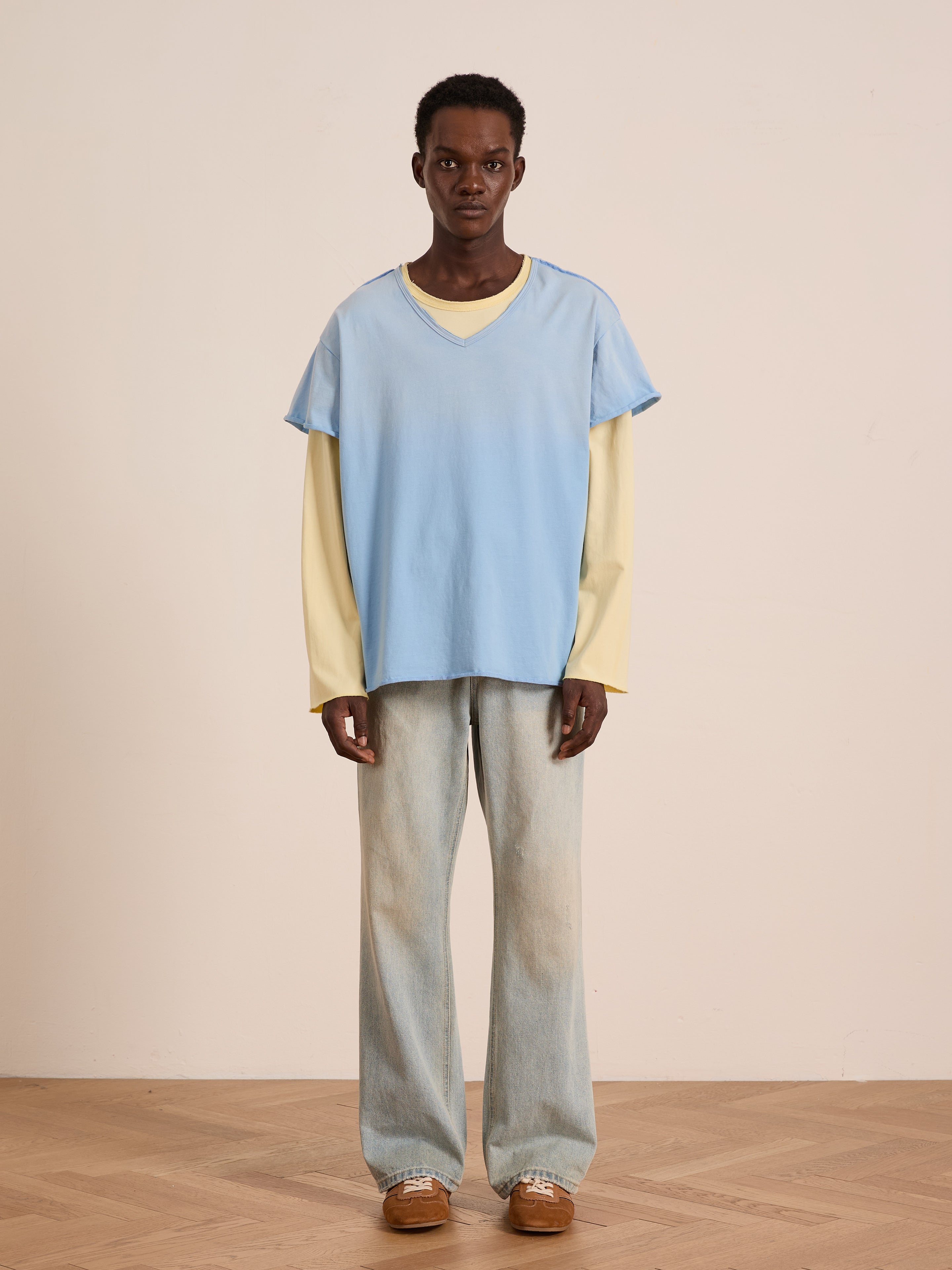 Indoors, a person wears the FOUND Double Layer LS Tee in light blue over a pale yellow long sleeve tee, paired with loose sunfade wash jeans and brown shoes against a plain, neutral background.