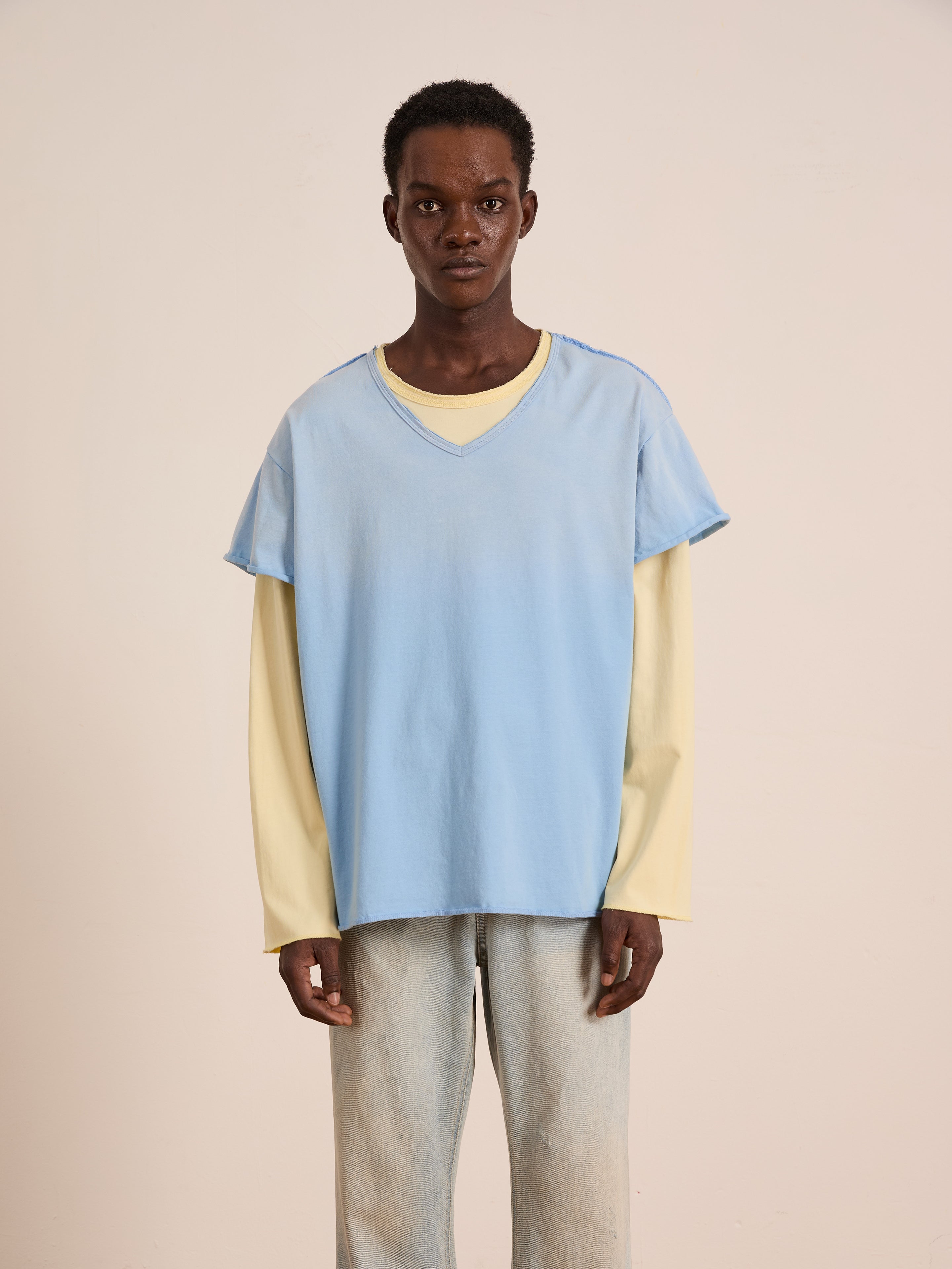 A person stands against a plain background, wearing FOUND's Double Layer LS Tee—a light blue short-sleeve top layered over a yellow unisex long sleeve—paired with light-wash jeans.