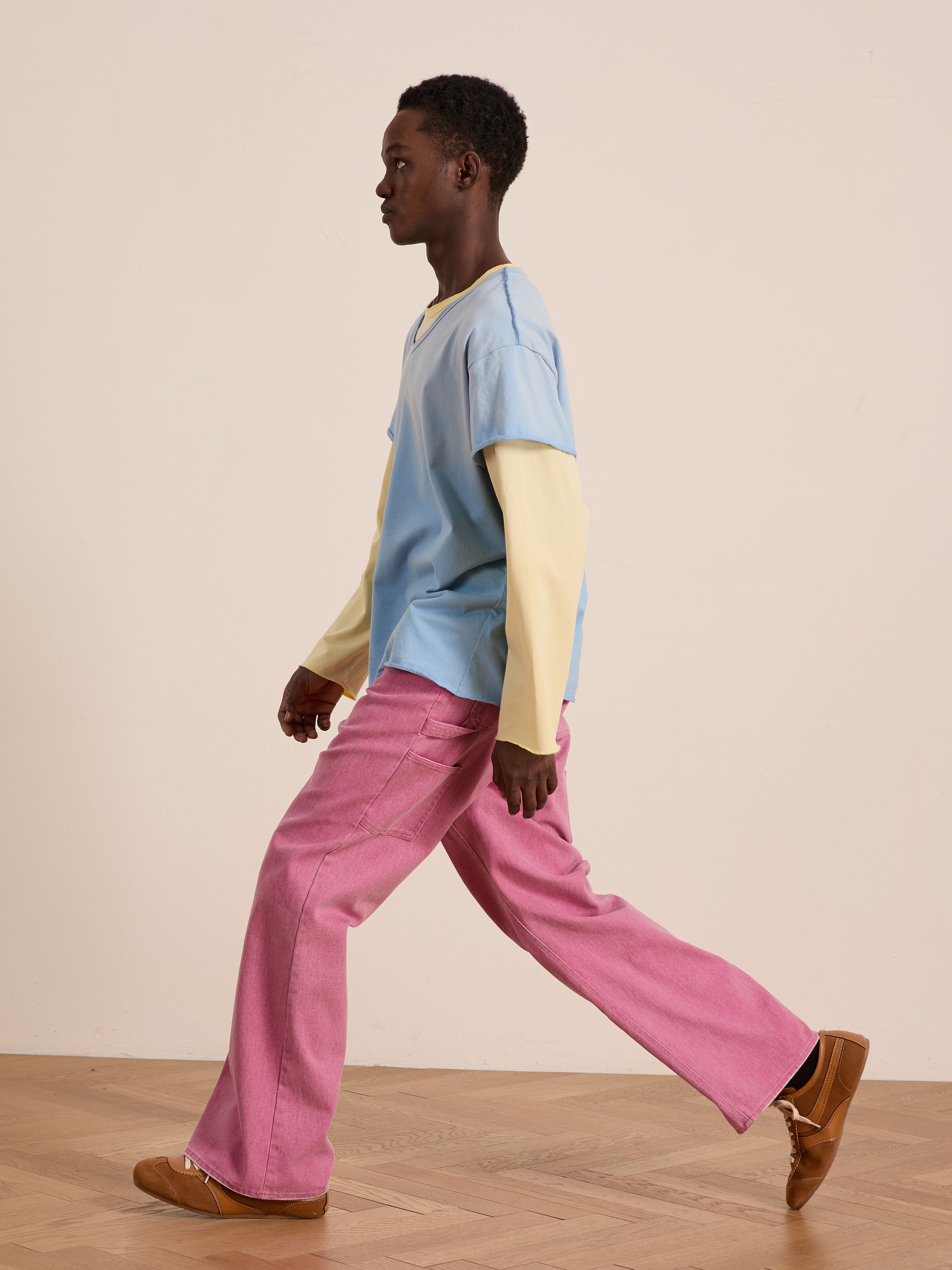 A person wears the FOUND Double Layer LS Tee—yellow long-sleeve under blue T-shirt—with pink pants and brown shoes, walking indoors against a plain light-colored wall.